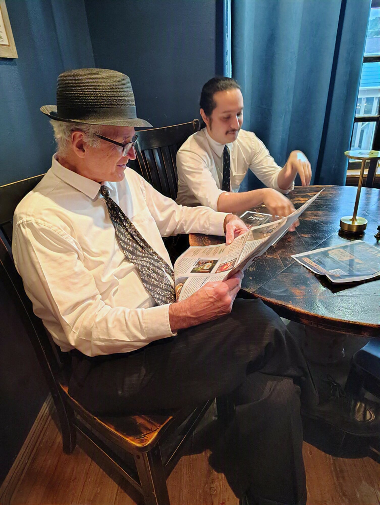Two men sit at a table reading newspapers, one wearing a hat and glasses, the other with a mustache, in a room with blue curtains.
