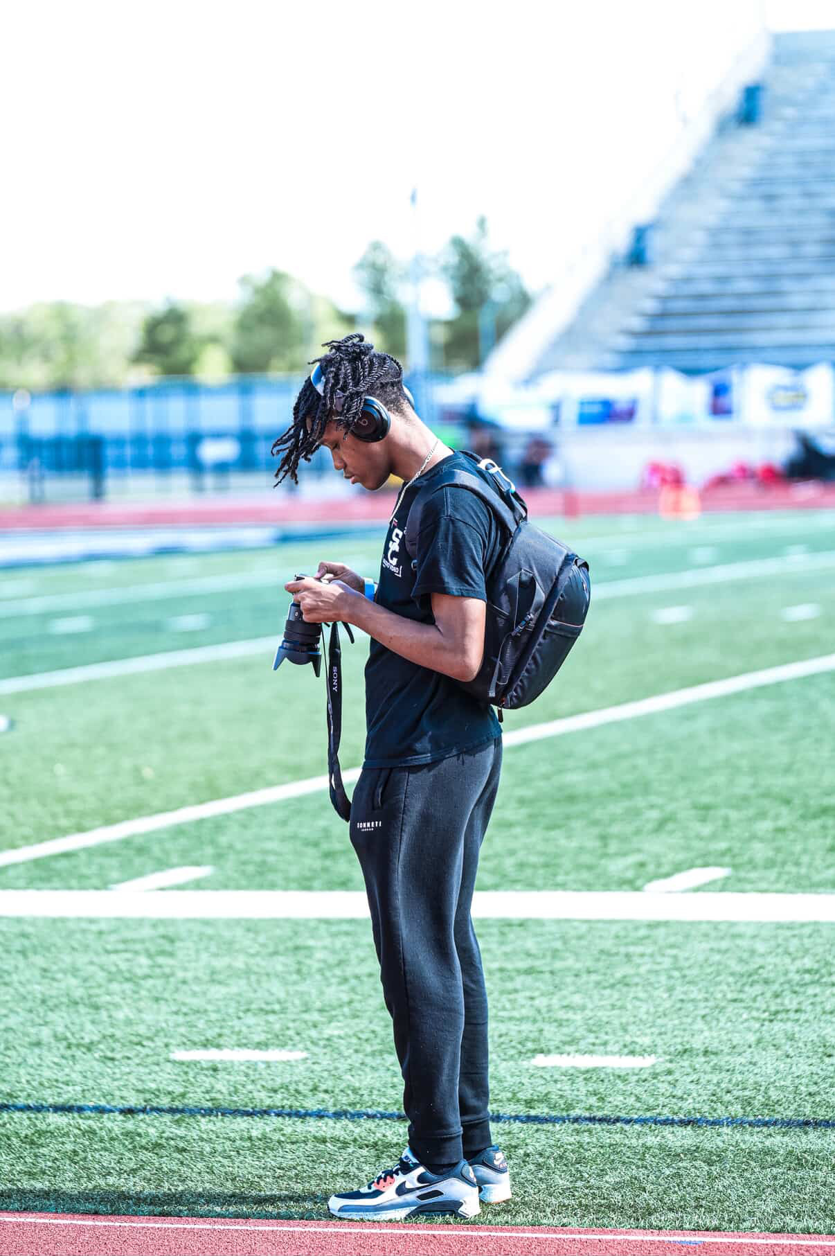 Person with dreadlocks looking at a camera on a sports field, wearing a backpack and athletic clothing.