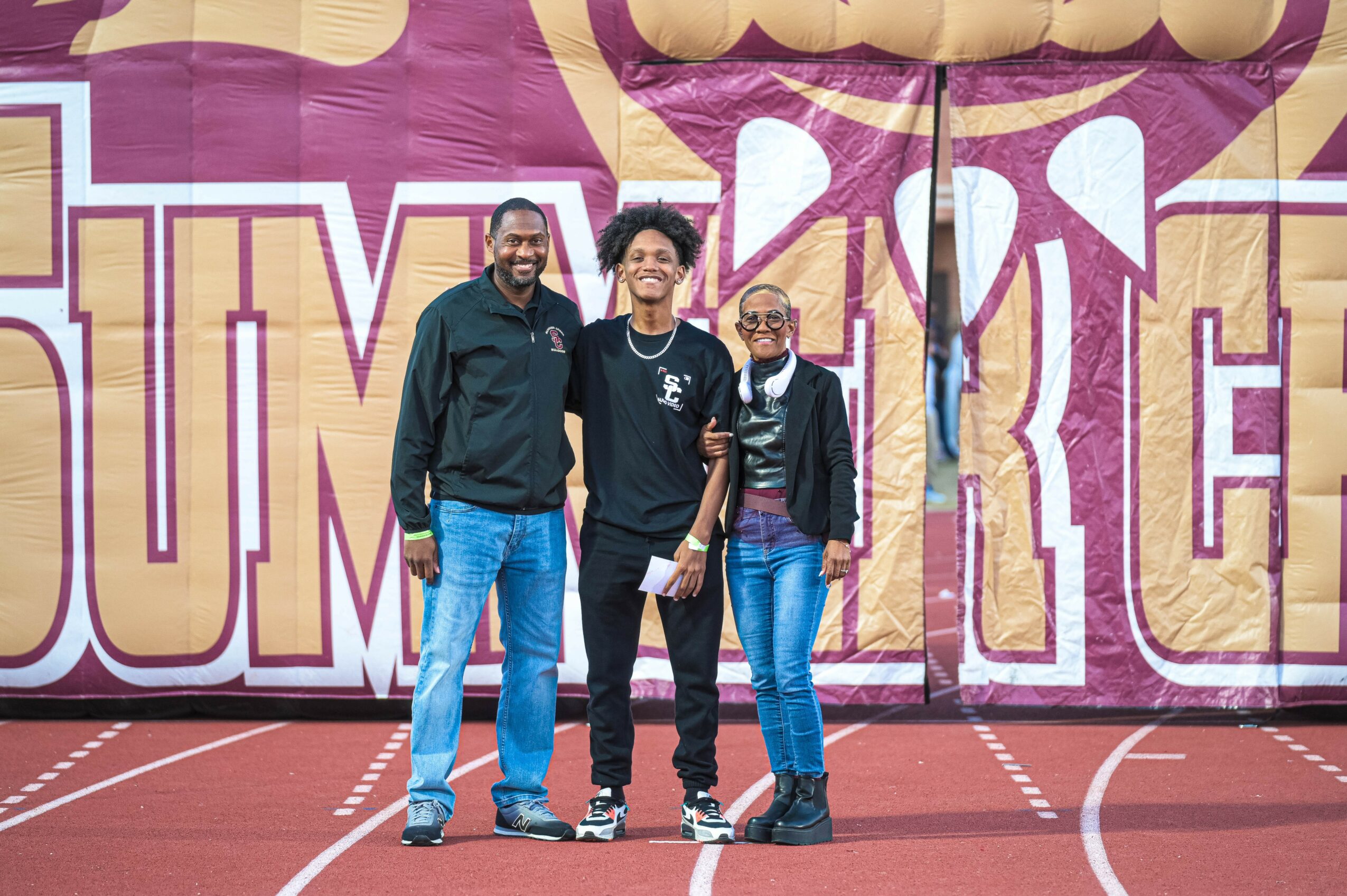Three people standing on a track in front of a large colorful mural, smiling, with arms around each other.
