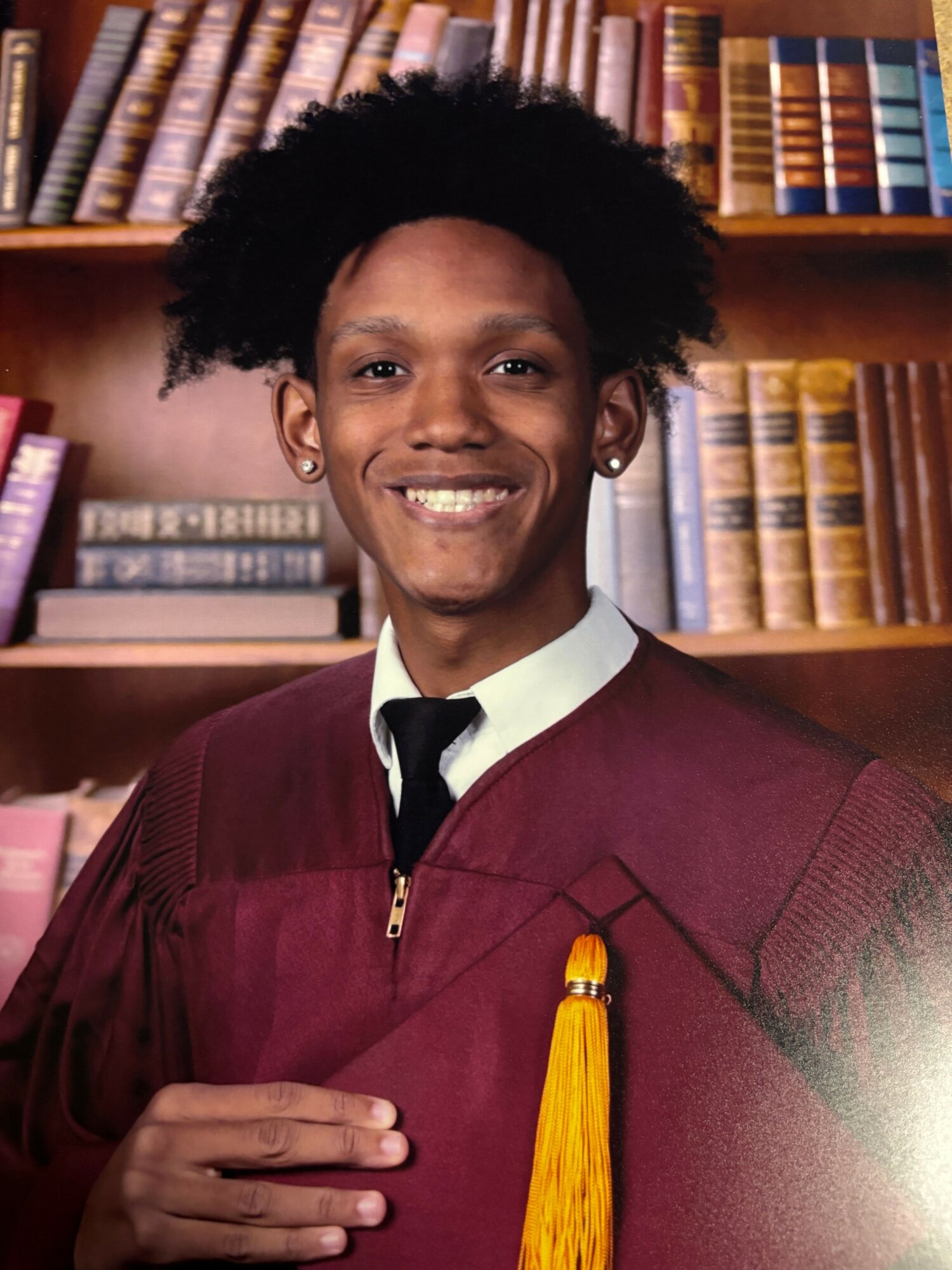 Person in graduation gown holding diploma, smiling, in library with bookshelves in background.