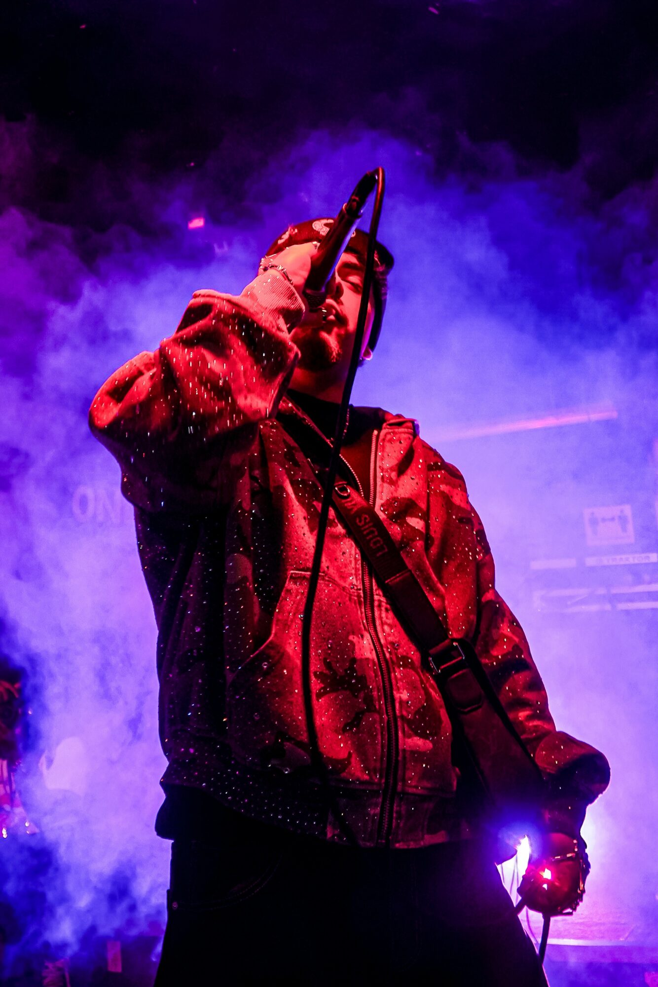 Person performing on stage with purple and blue lighting, holding a microphone, wearing a red jacket, surrounded by smoke.