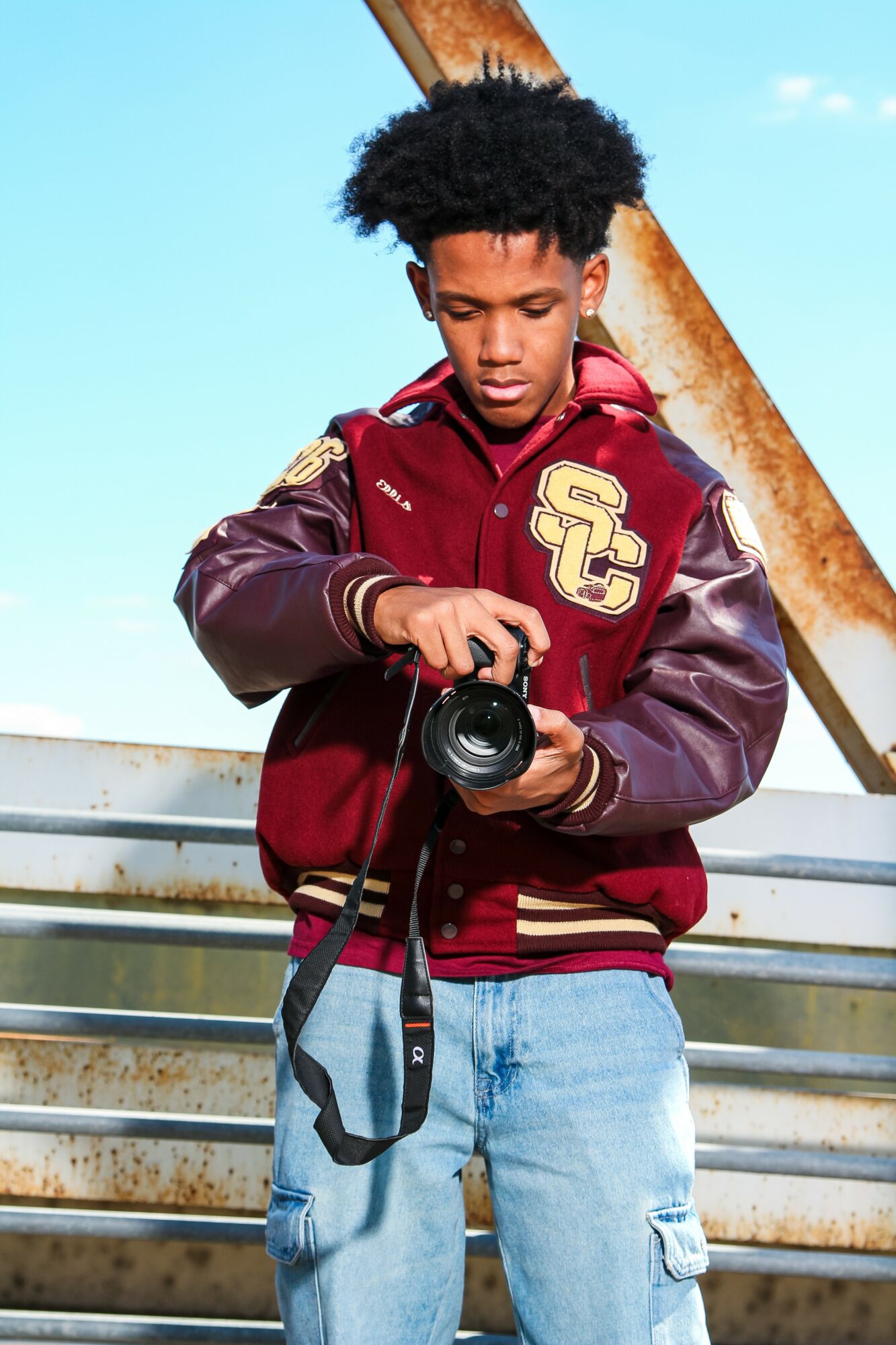 Young person with curly hair looking at camera, holding a camera, wearing a red and brown varsity jacket, standing outdoors.