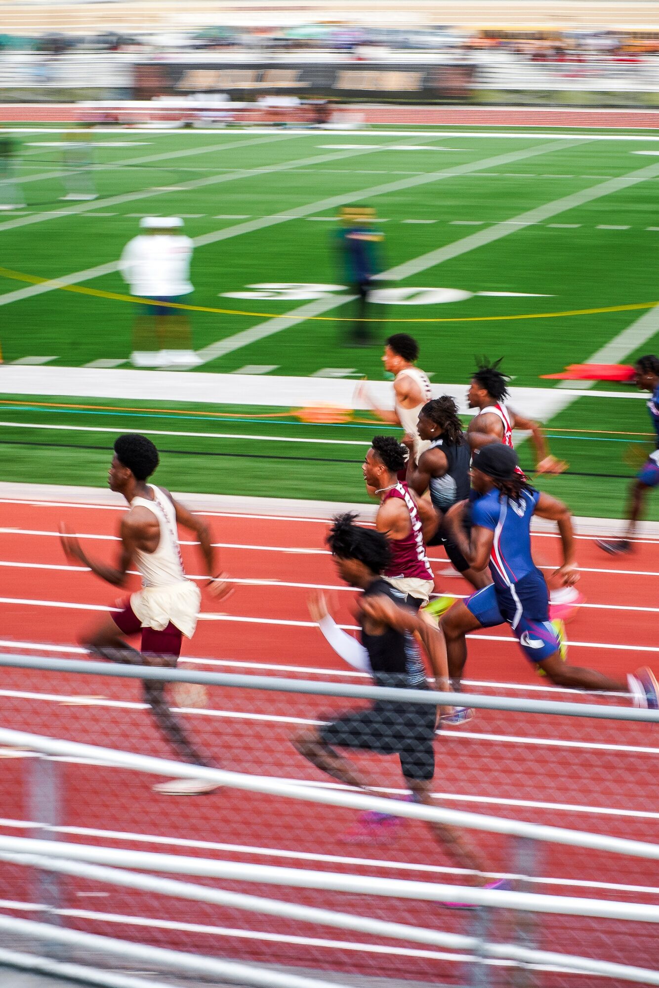 Group of runners on a red track during a race, with a green field and blurred spectators in the background.