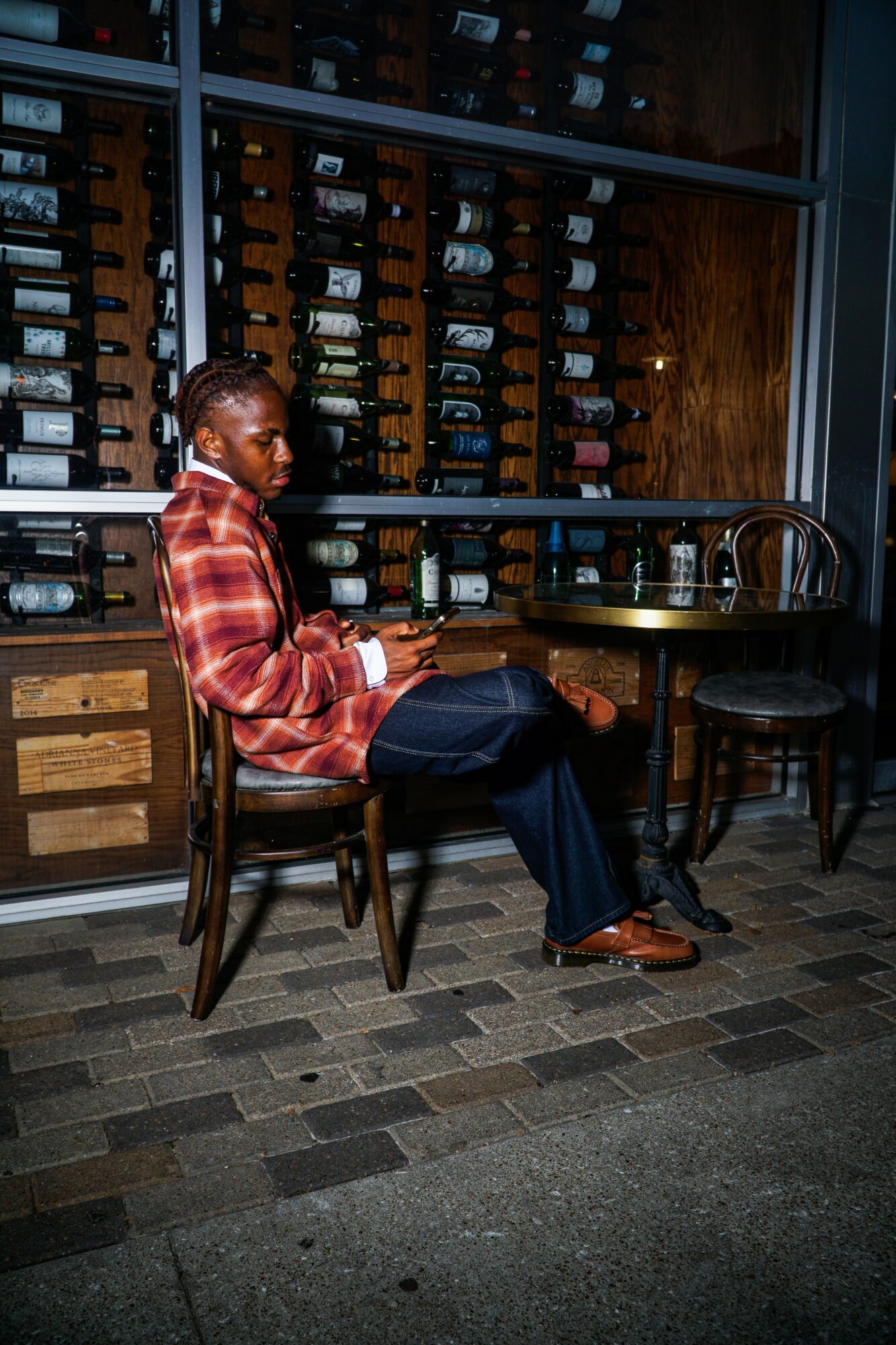 Person sitting at a table in a wine cellar with wine bottles on shelves behind, wearing a red plaid shirt and dark pants.