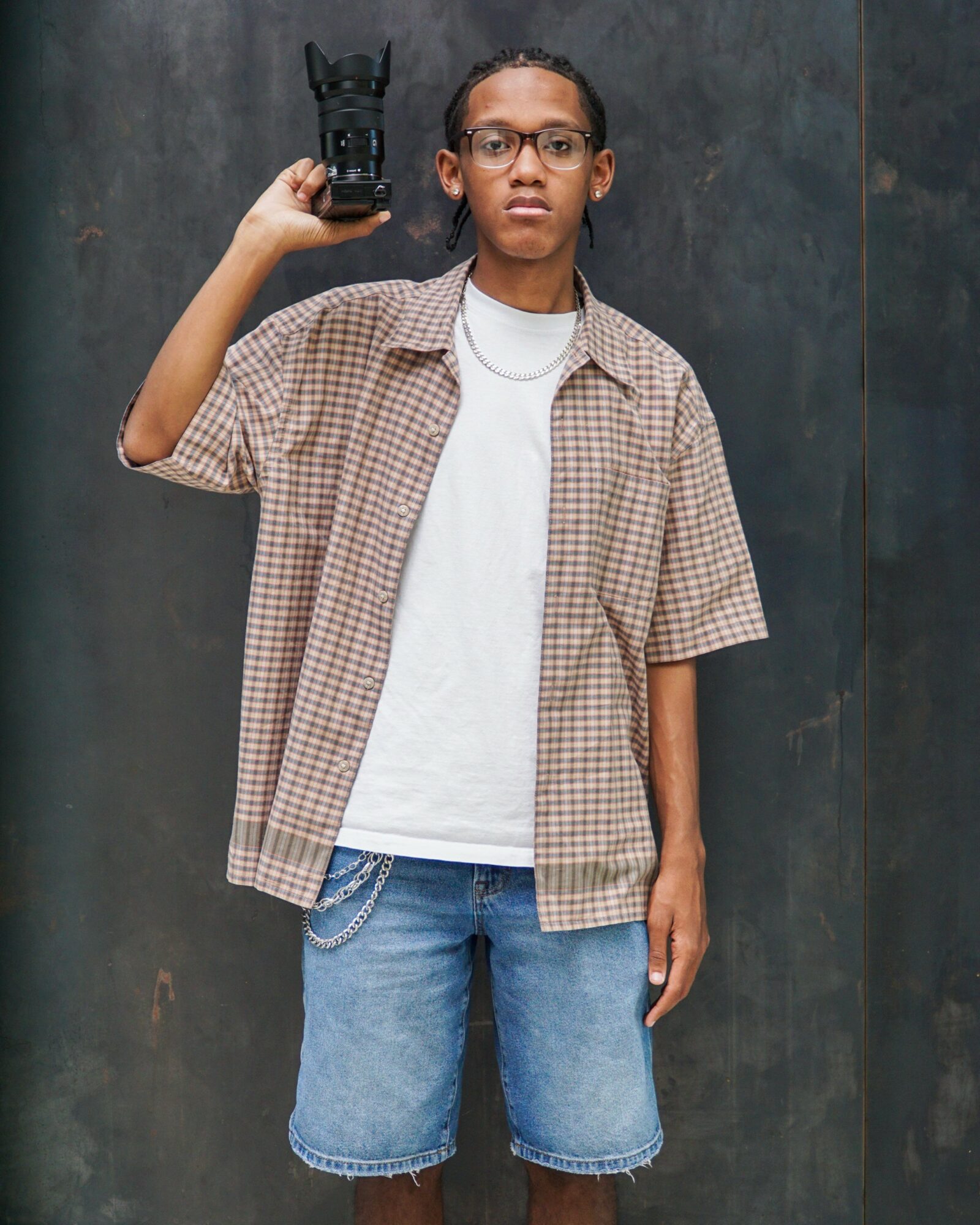 Person holding a camera, wearing glasses, a checkered shirt, white t-shirt, and denim shorts, standing against a dark wall.