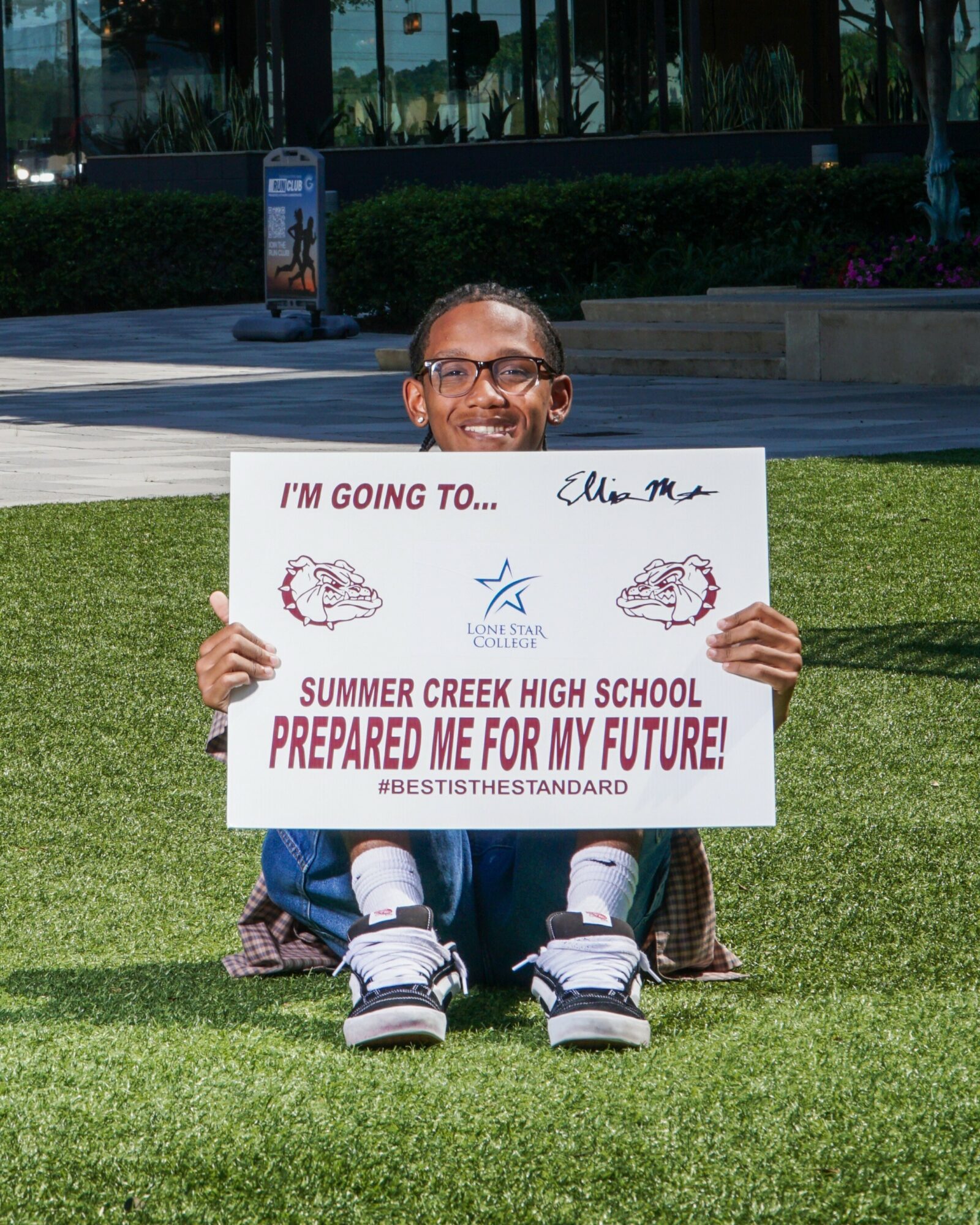 Child sitting on grass holding a sign with text and signatures, outdoors with trees and a sidewalk in the background.