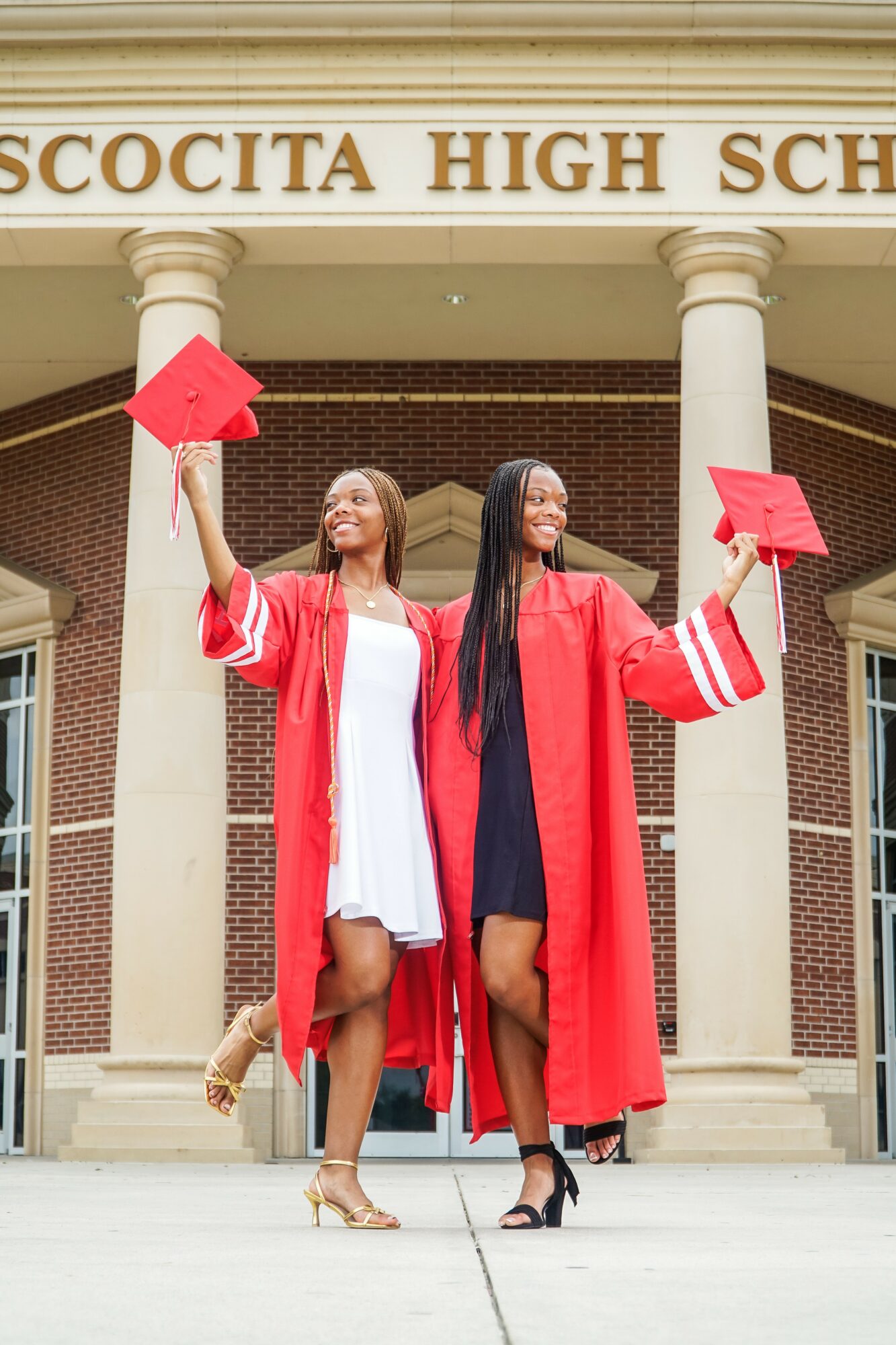Two graduates in red gowns and caps celebrate in front of a high school building, holding their caps in the air.