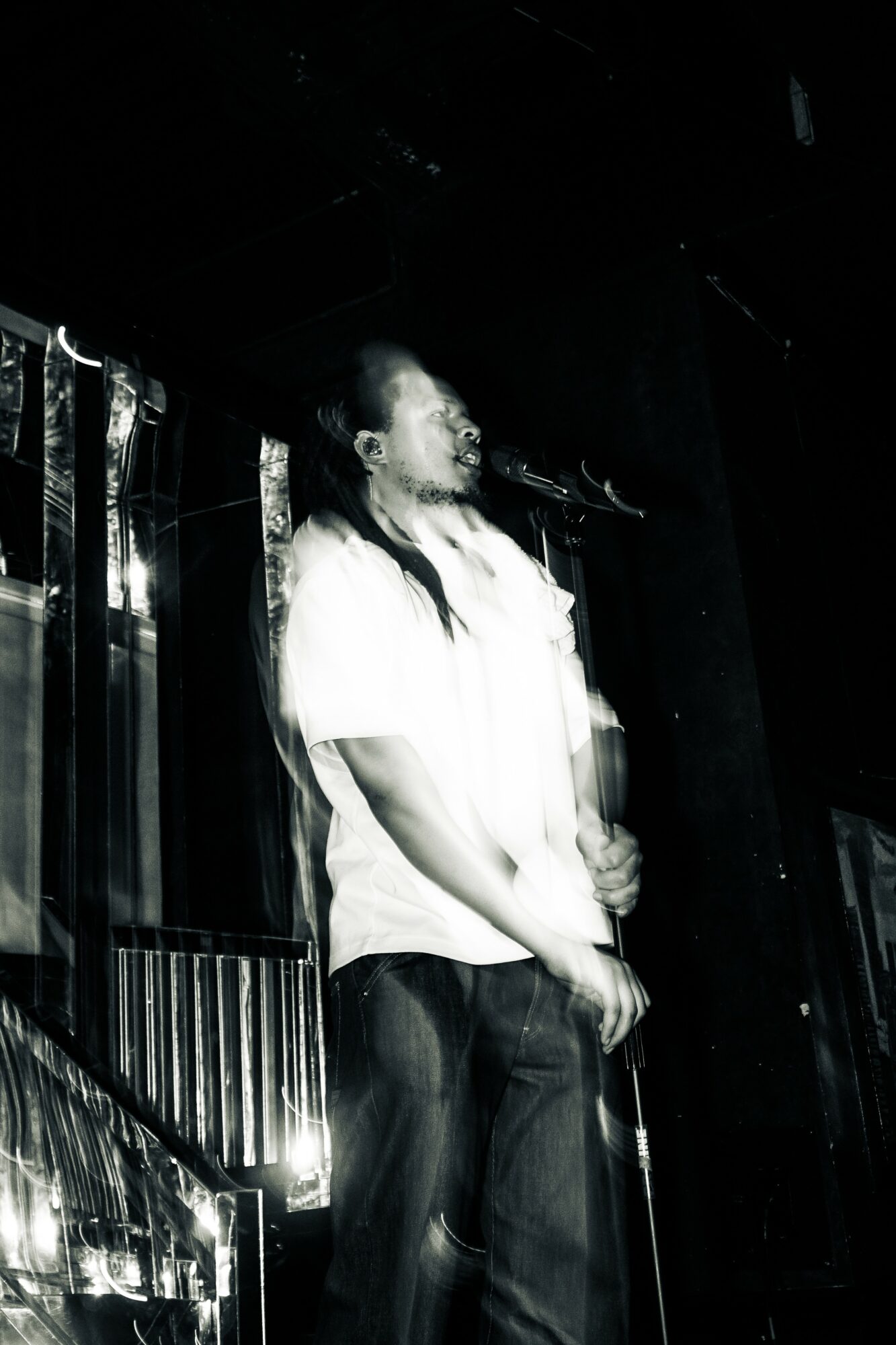 Person standing on stage with microphone, blurred motion, dark background, stage equipment visible.