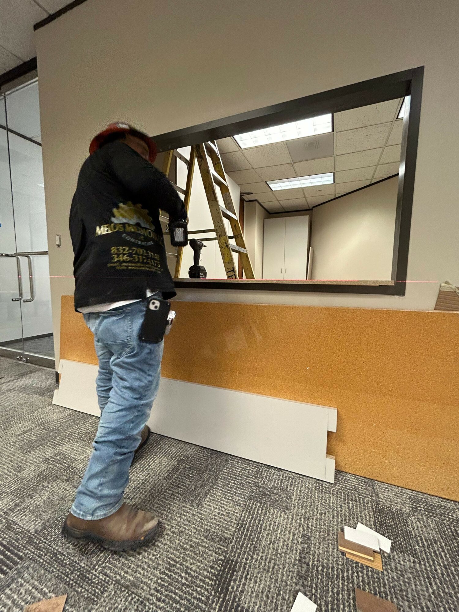 Person standing in an office space, looking at a large mirror, with a carpeted floor and office furniture nearby.