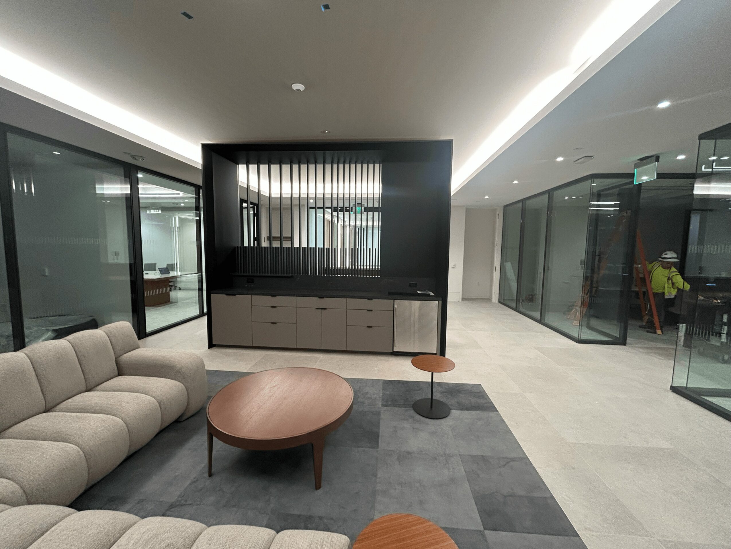 Modern lobby with beige sofa, round coffee table, and a black partition with drawers, glass walls, and a hallway.