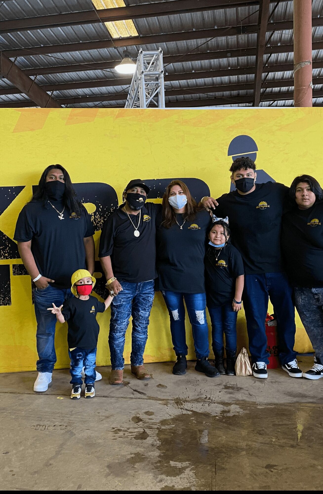 Group of people and children standing in front of a yellow wall with a vehicle behind them, indoors with a high ceiling.