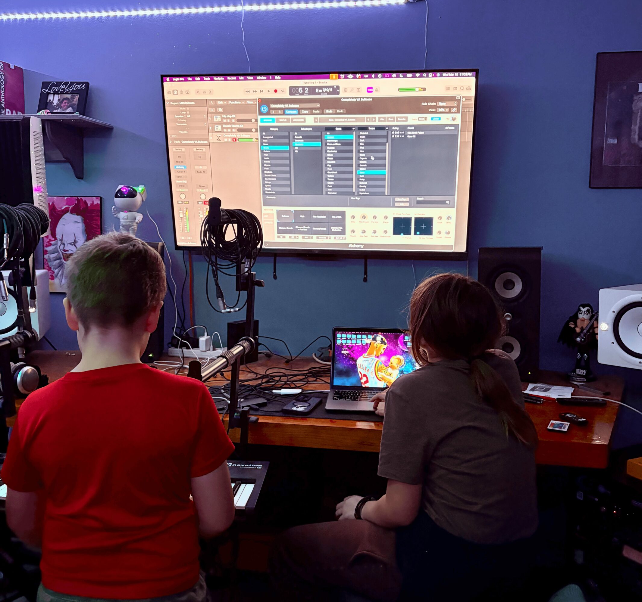 Two children in a music studio with a large screen displaying music software, one standing and one sitting at a desk.