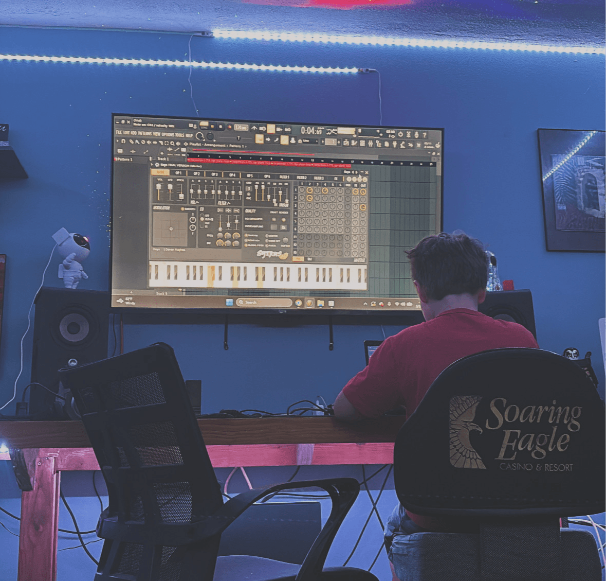 Person with dark hair and red shirt sitting in front of a large screen displaying music production software in a blue-lit room.