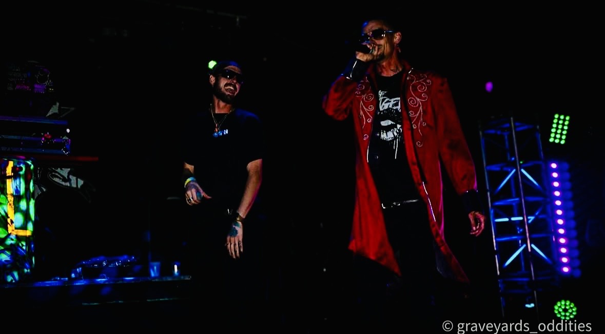 Two people on stage with colorful lighting and equipment in the background, one holding a microphone, performing.