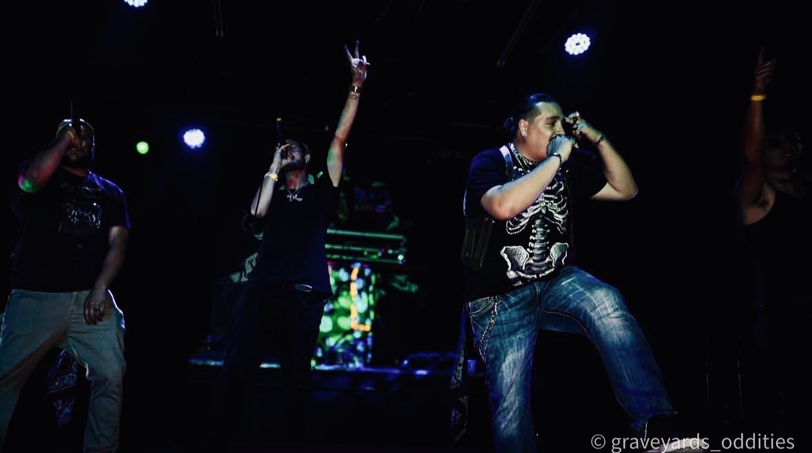 Group of four people performing on stage with dark background and colorful lights, one person singing into a microphone.