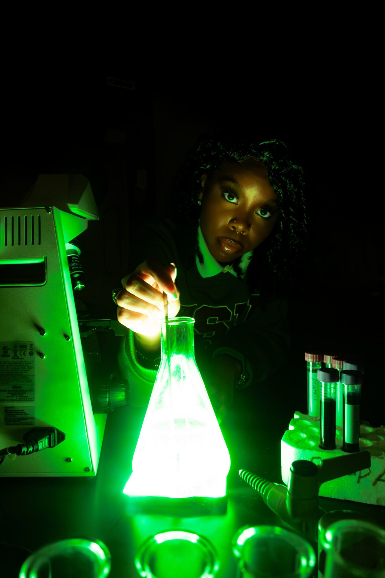 Child with curly hair in a dark room, illuminated by green glow from laboratory equipment, holding a glowing flask.