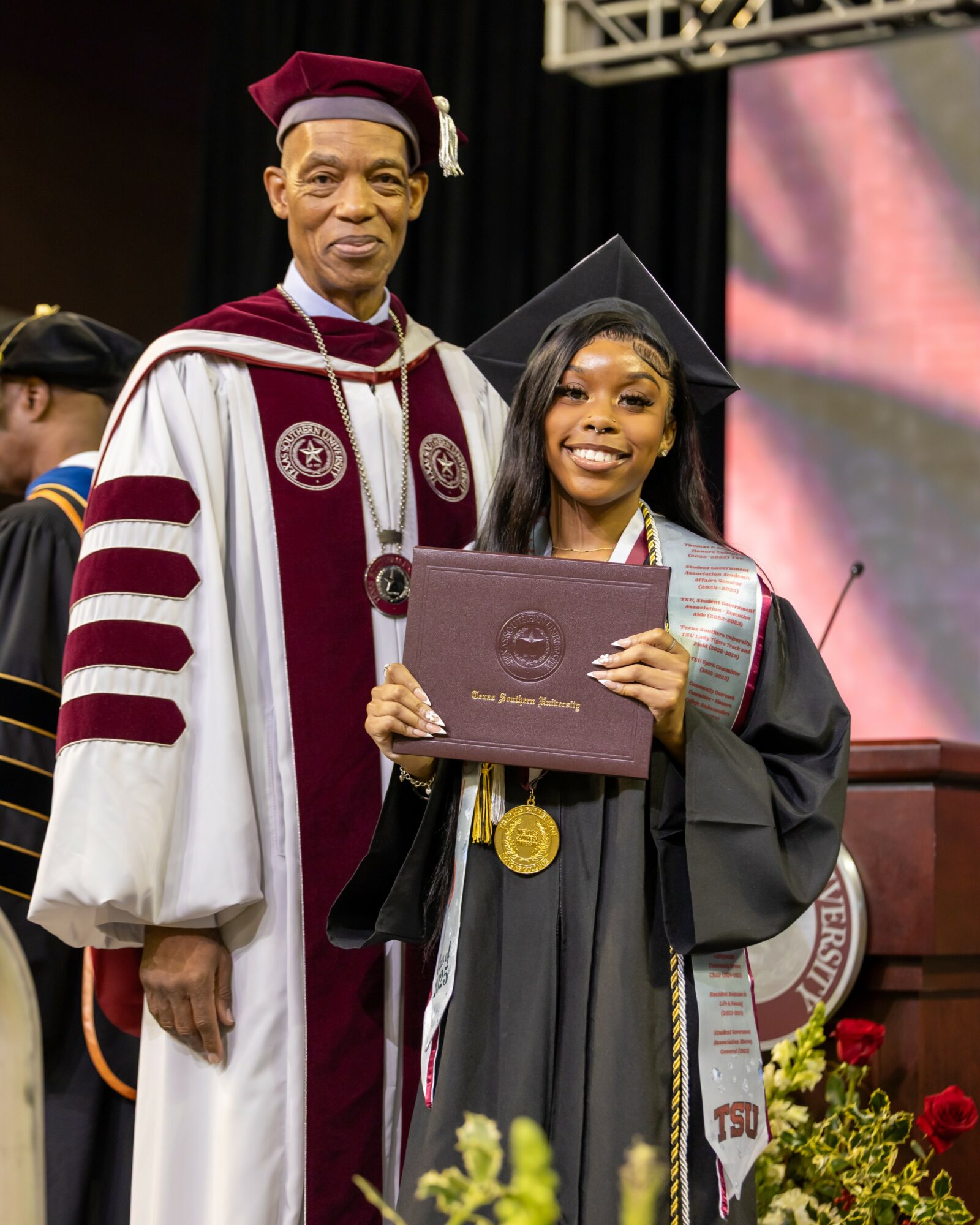 Young woman in graduation cap and gown holding diploma, smiling, standing next to an older man in academic regalia, on stage.
