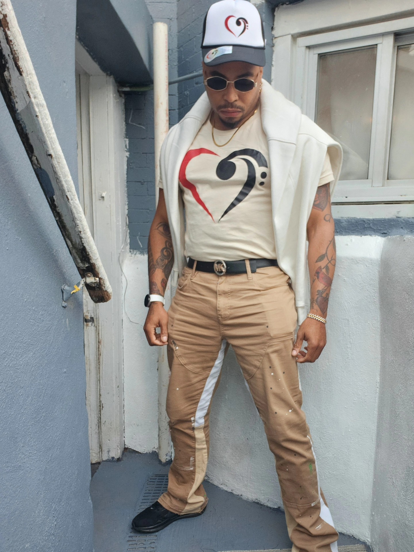 Man wearing sunglasses, a cap, a graphic t-shirt, beige pants, and black shoes, standing outdoors near a building.