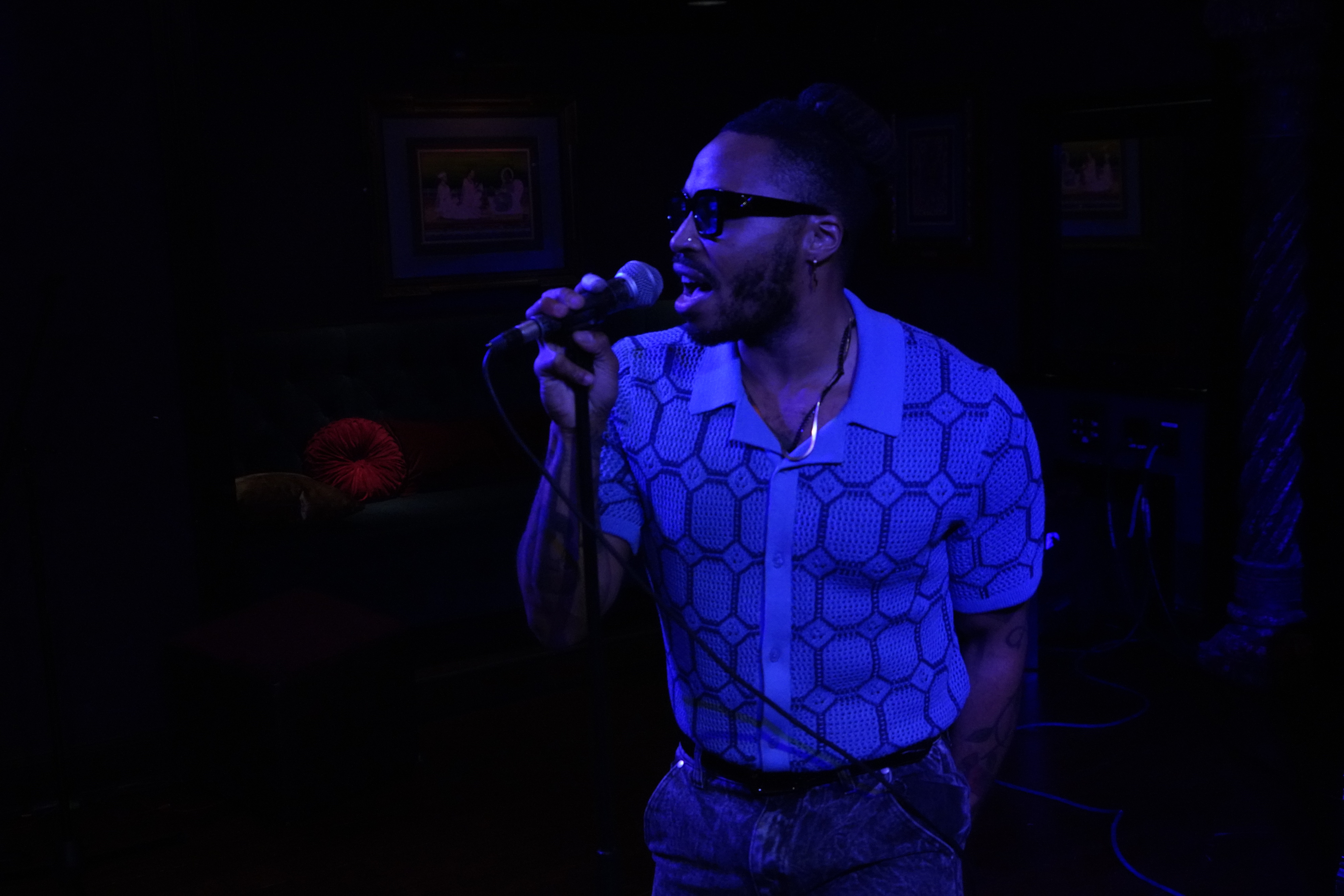 Man singing into a microphone on stage, wearing glasses and a patterned shirt, with dark background and dim lighting.