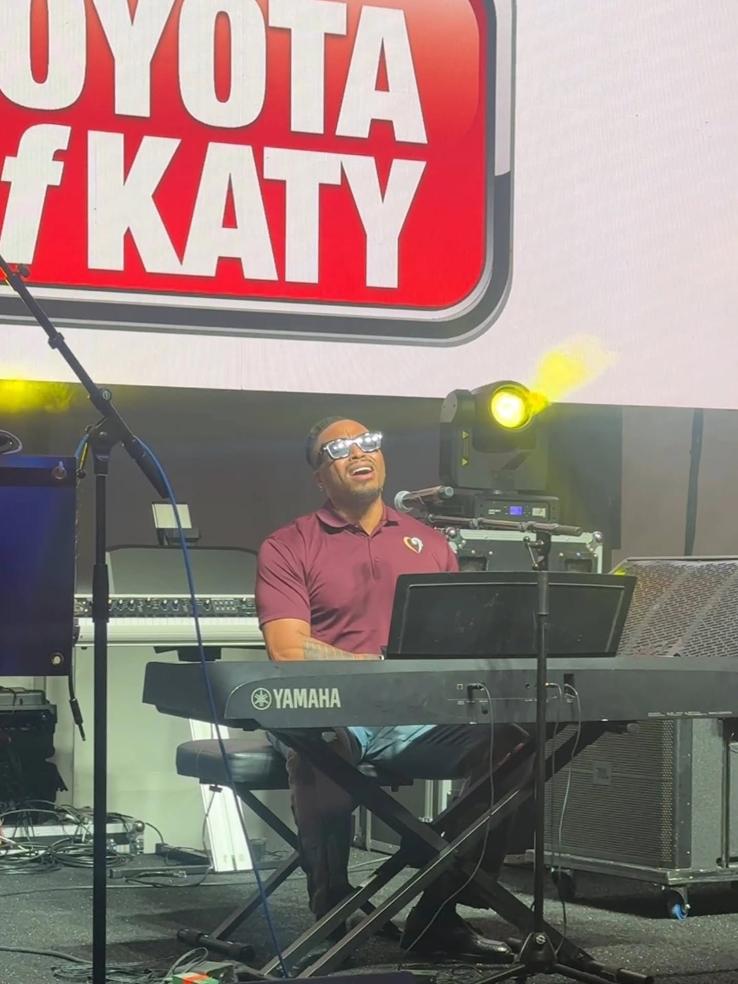 Person playing Yamaha keyboard on stage with a sign reading 'Toyota of Katy' above, stage lights shining, person wearing glasses and maroon shirt.