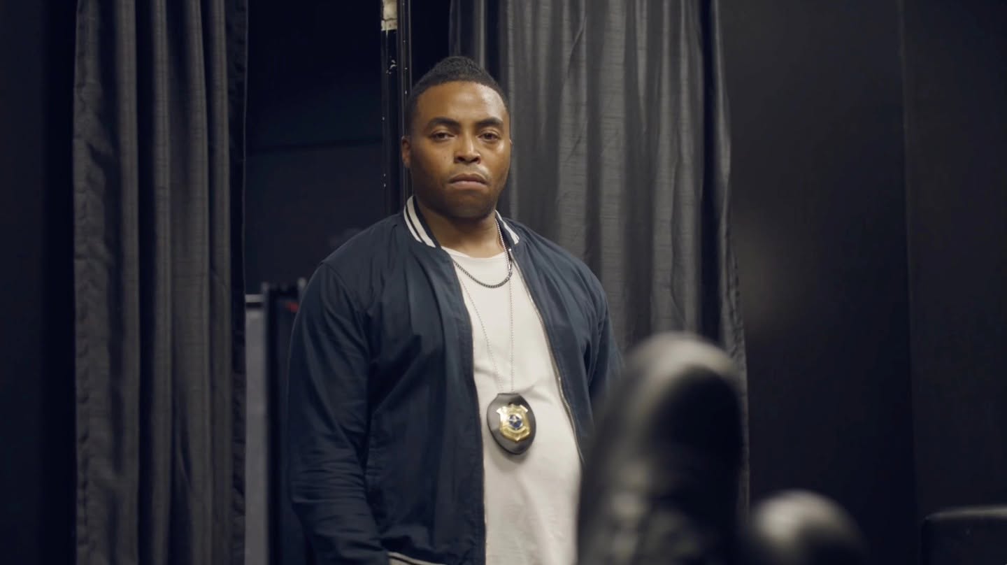 A man stands in front of dark curtains, wearing a white shirt, black jacket, and a badge around his neck.