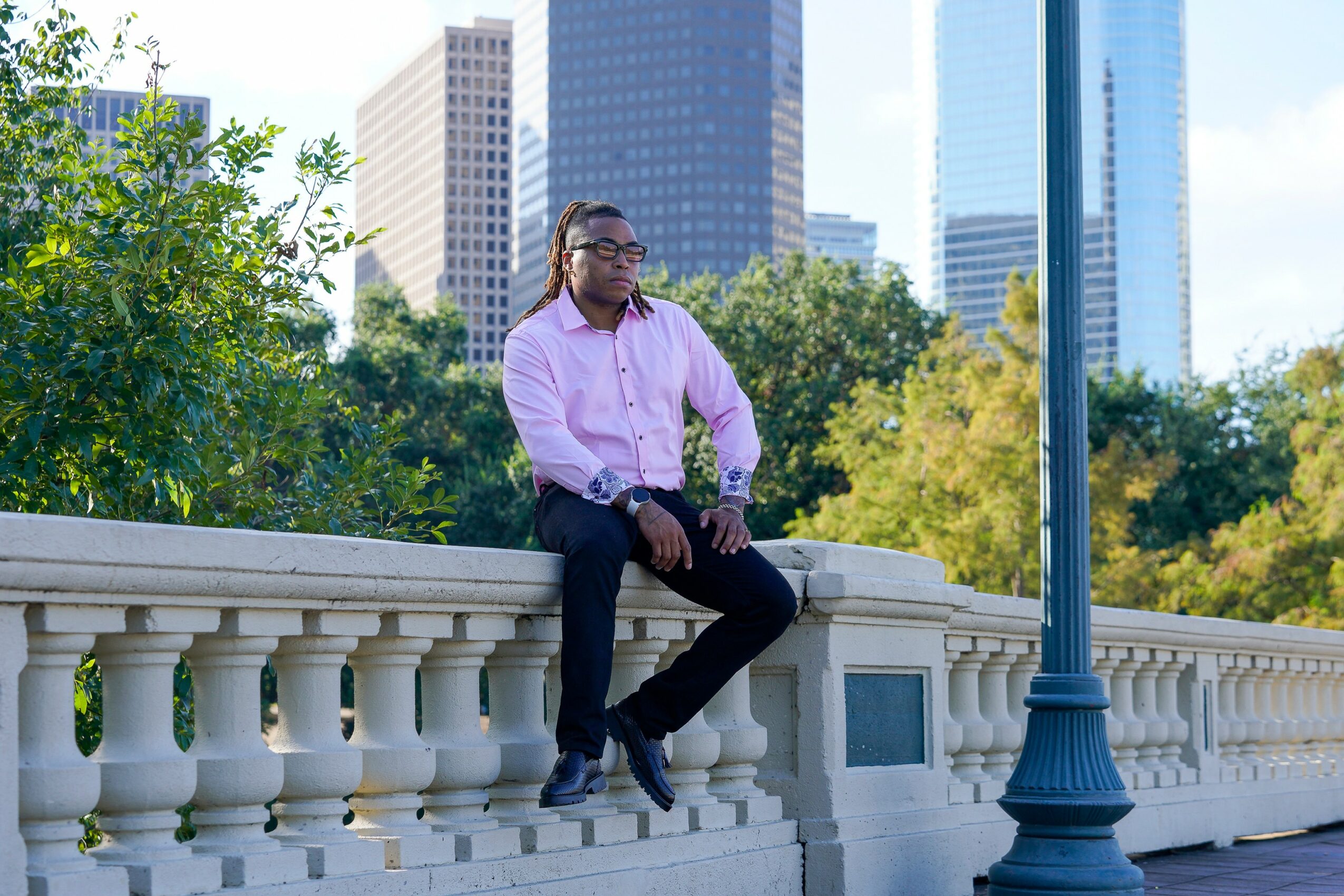Person sitting on a white stone railing in an urban park with tall buildings in the background.