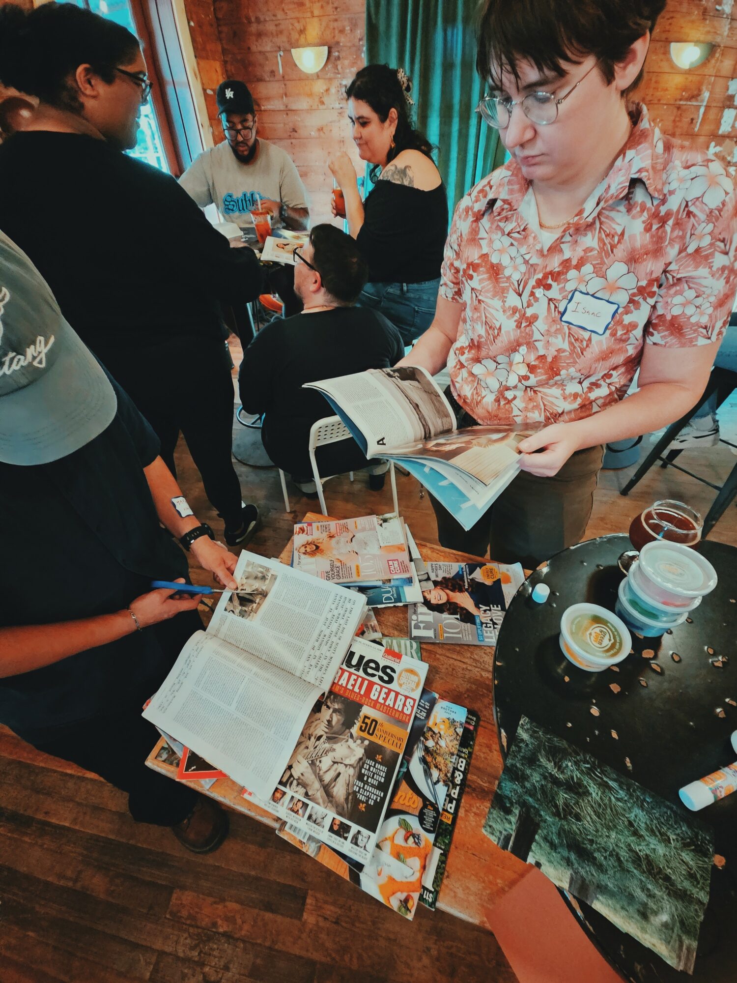 Group of people gathered around a table with magazines, books, and cups, engaging in conversation and browsing materials.