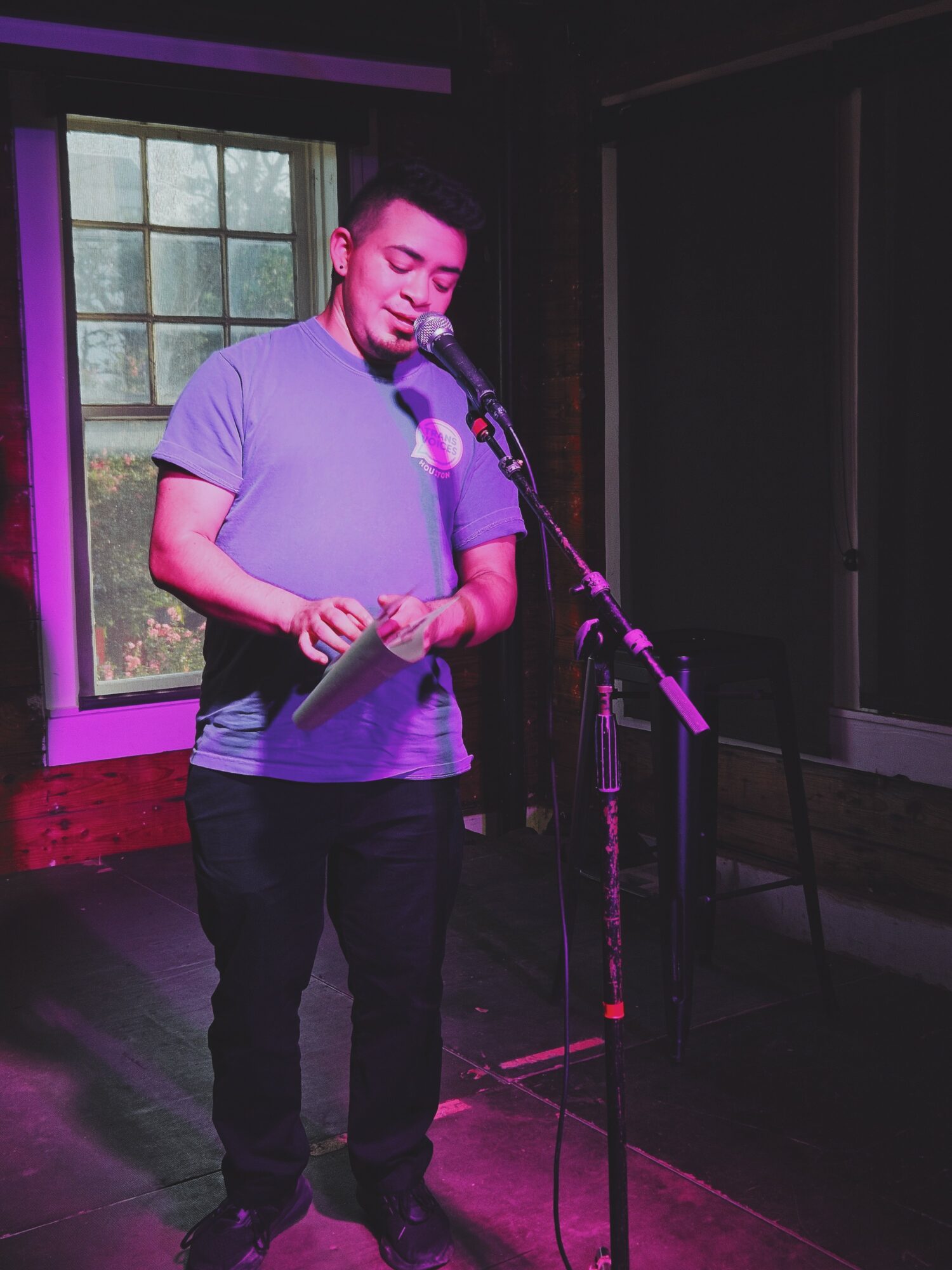 Person standing on stage, holding a paper, speaking into a microphone, with a window in the background.