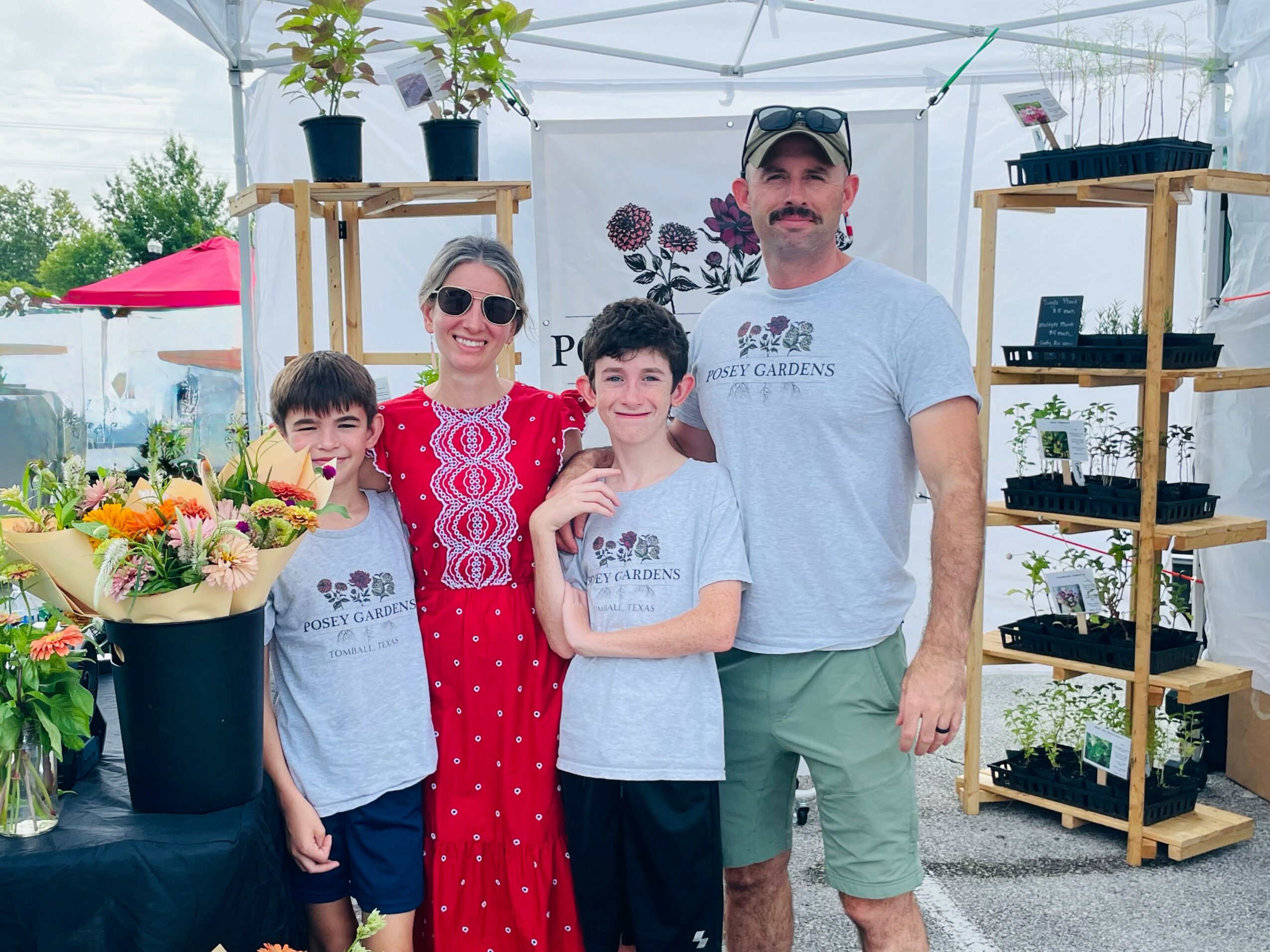 Family of four standing outdoors at a plant nursery, smiling, with shelves of plants behind them.