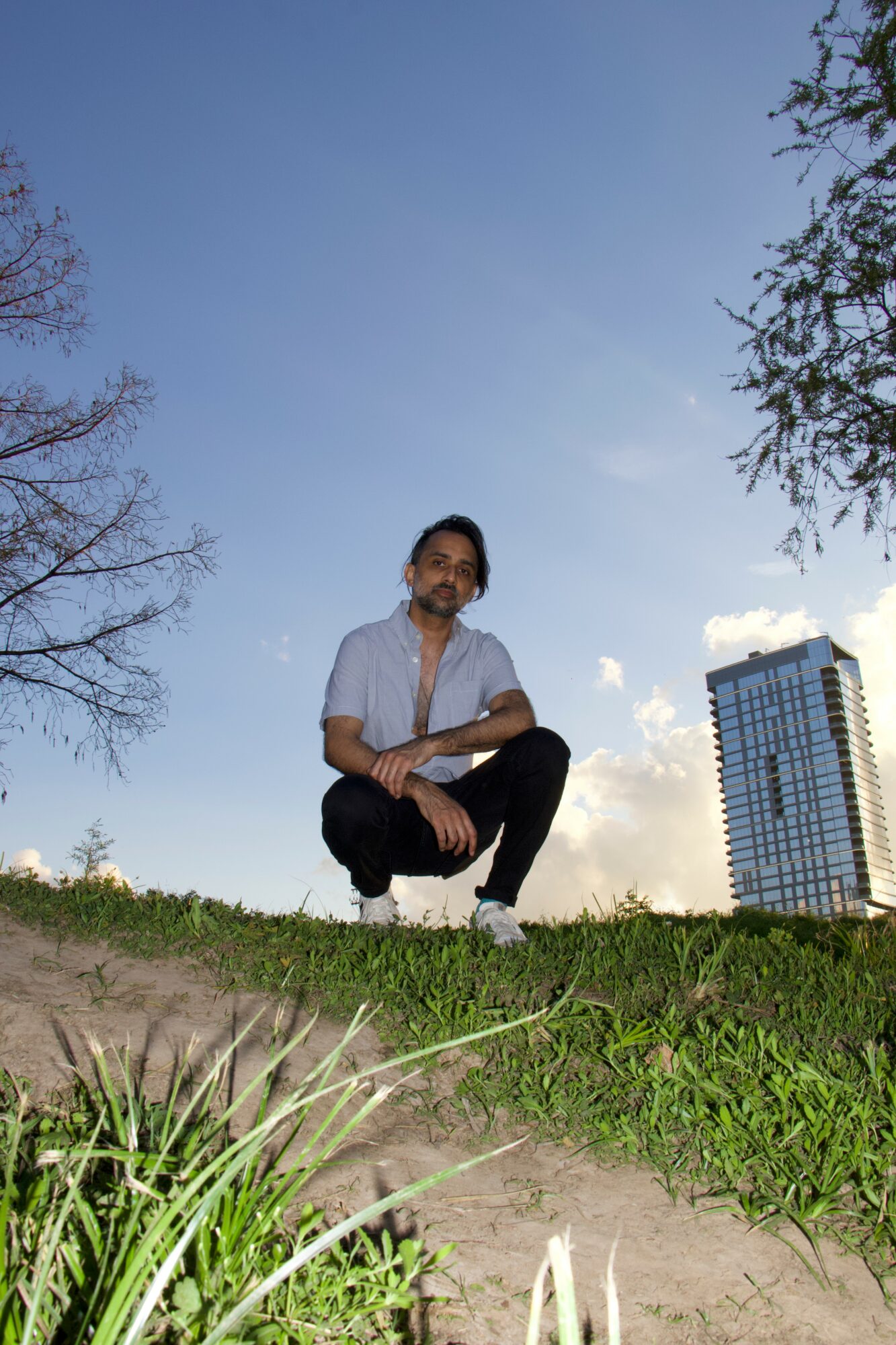 Person crouching on grass with a city building and trees in the background under a clear blue sky.