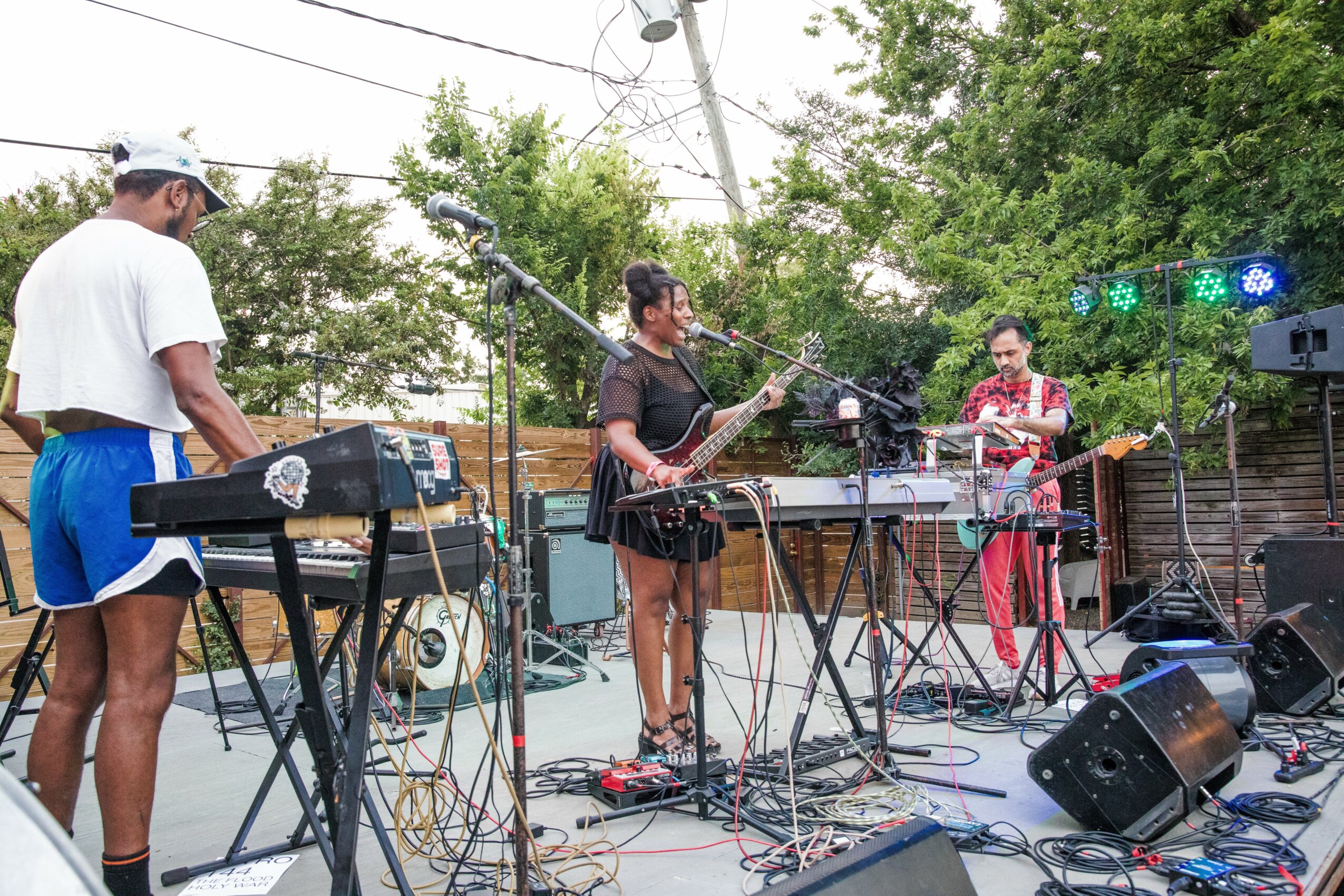 Three musicians perform outdoors with electronic instruments and microphones, surrounded by trees and equipment, under stage lighting.