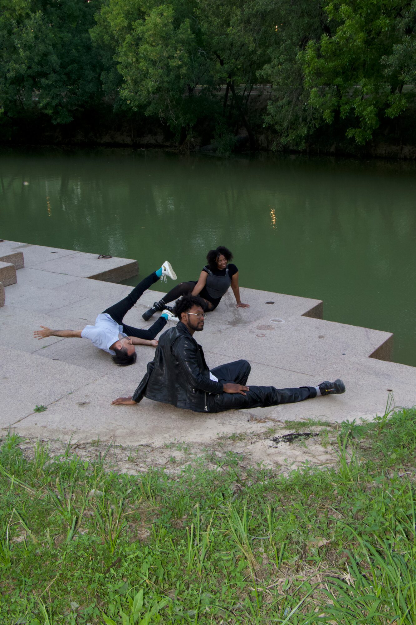 Three people sitting and lying on concrete steps by a river, with trees in the background.