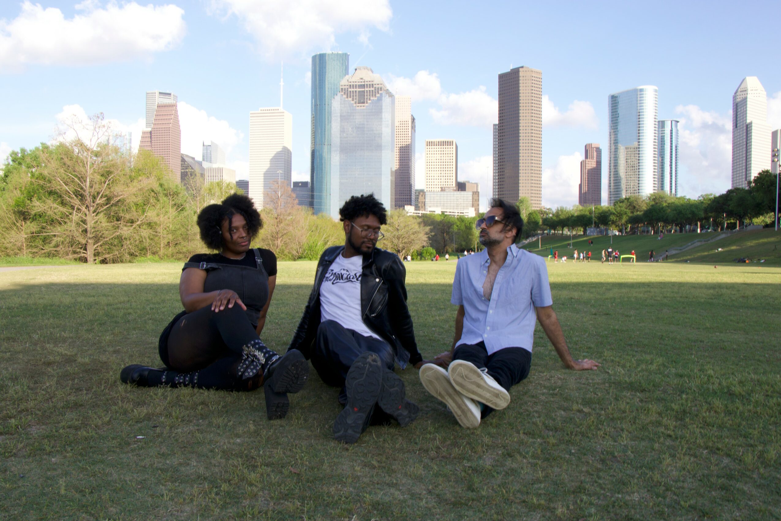 Three people sitting on grass with city skyline in background, trees, and blue sky with clouds.