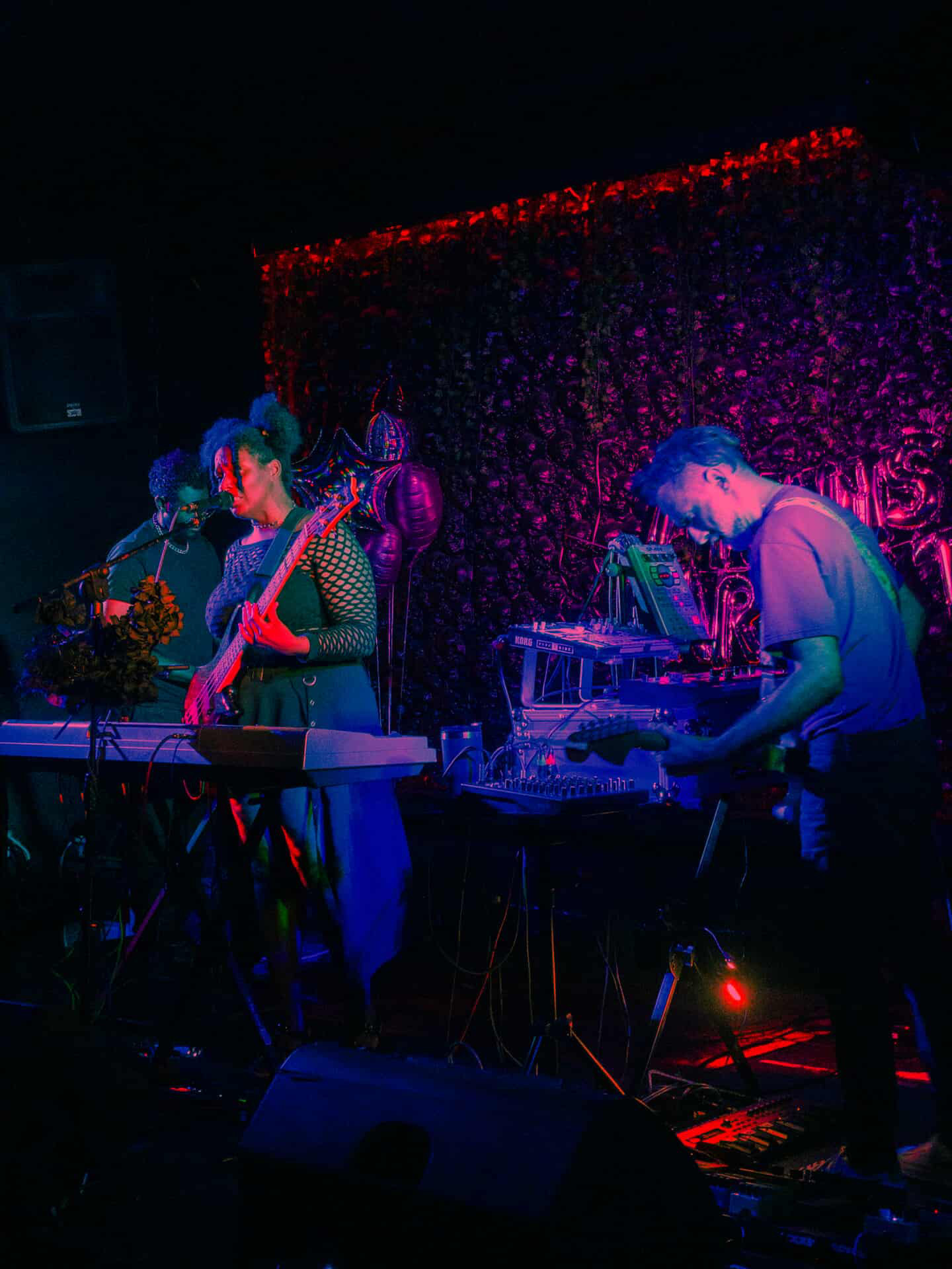 Musicians perform on stage with colorful lighting and a glittery backdrop, playing keyboards and electronic instruments.