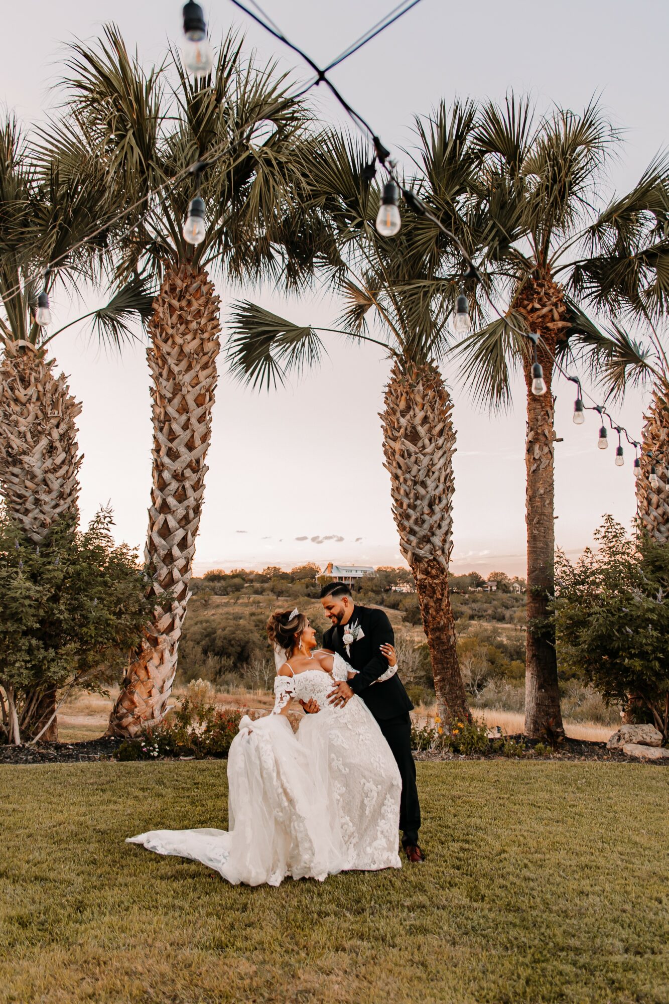 A bride and groom dance outdoors under palm trees with string lights, during sunset or dusk, on a grassy area.