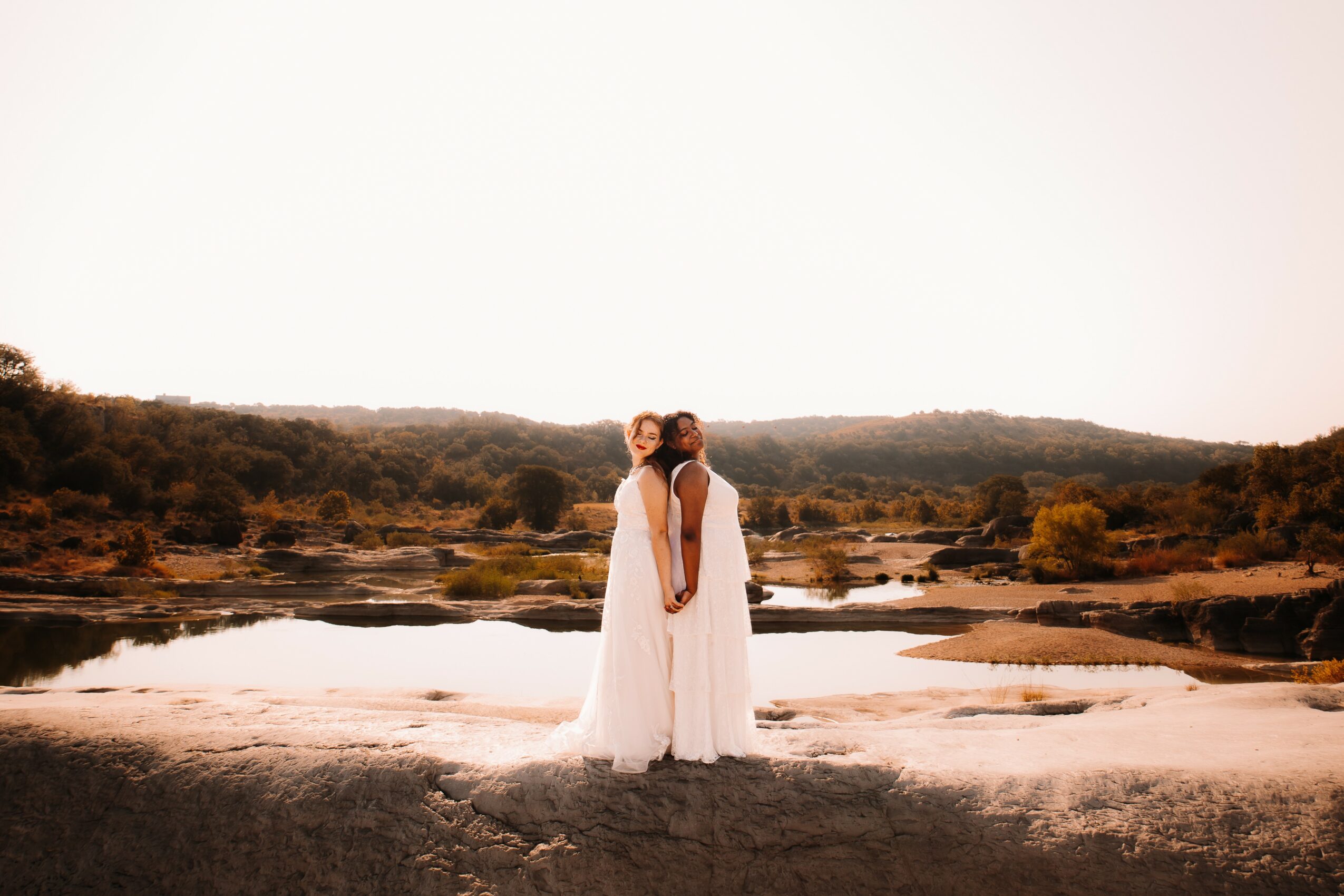 Two women in white dresses stand close together near a river, with a landscape of trees and hills in the background.