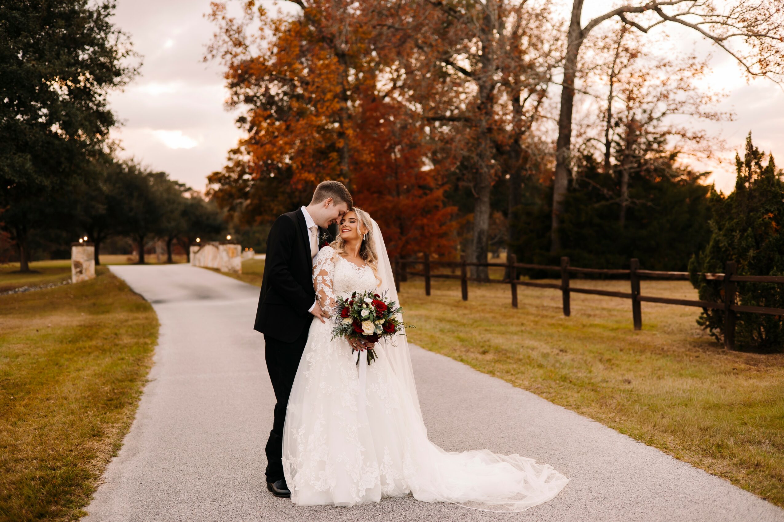 A bride and groom embrace on a winding path outdoors during autumn, with trees and a fence in the background.