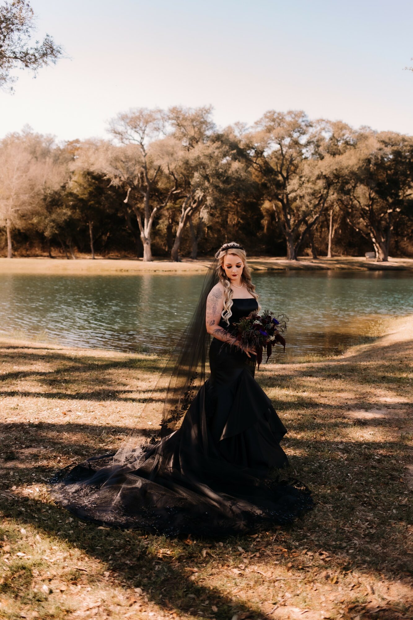 Woman in a black dress with a veil holding a bouquet stands outdoors near a lake with trees in the background.