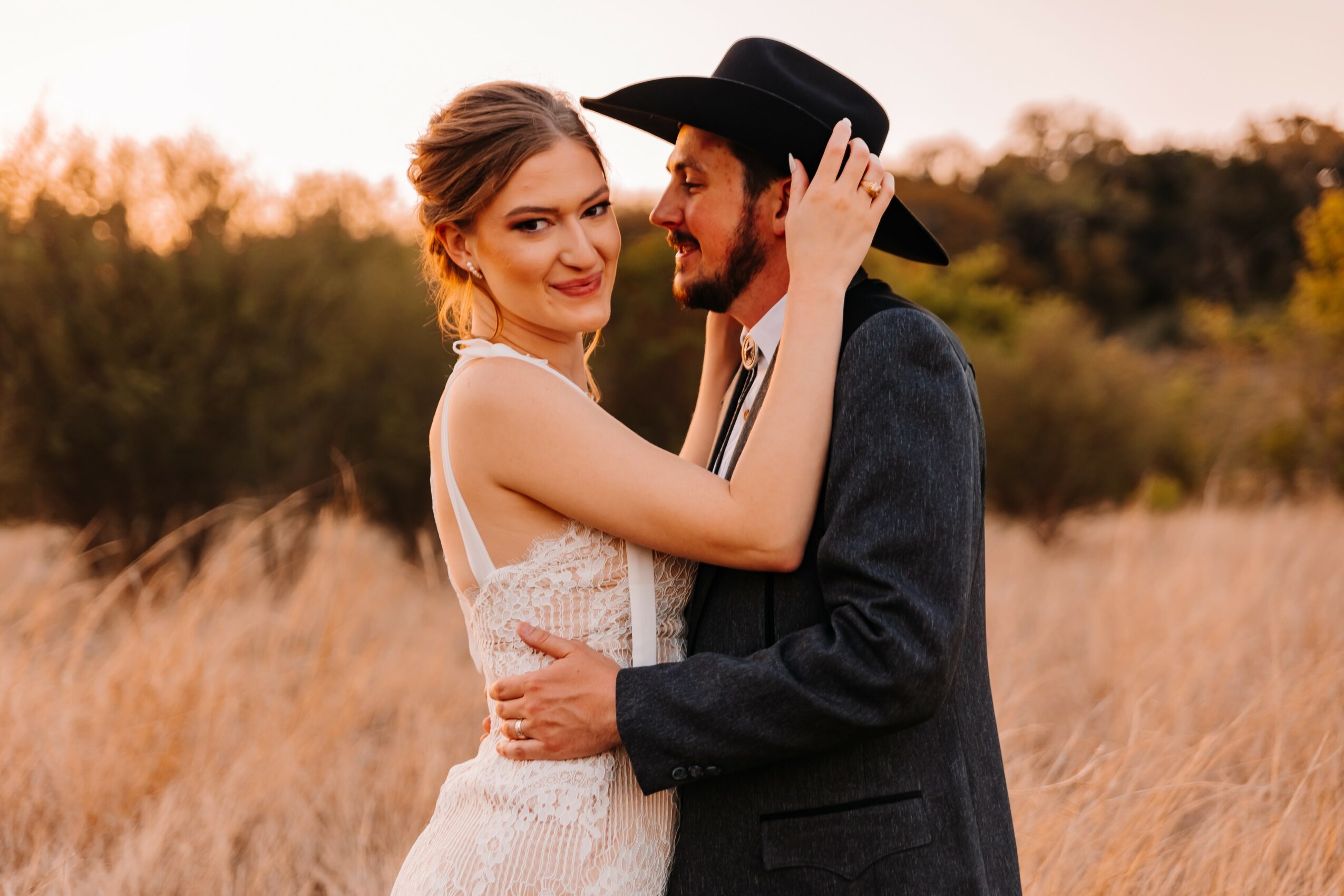A woman and man embrace outdoors during sunset, woman touching man's face, both smiling, field and trees in background.