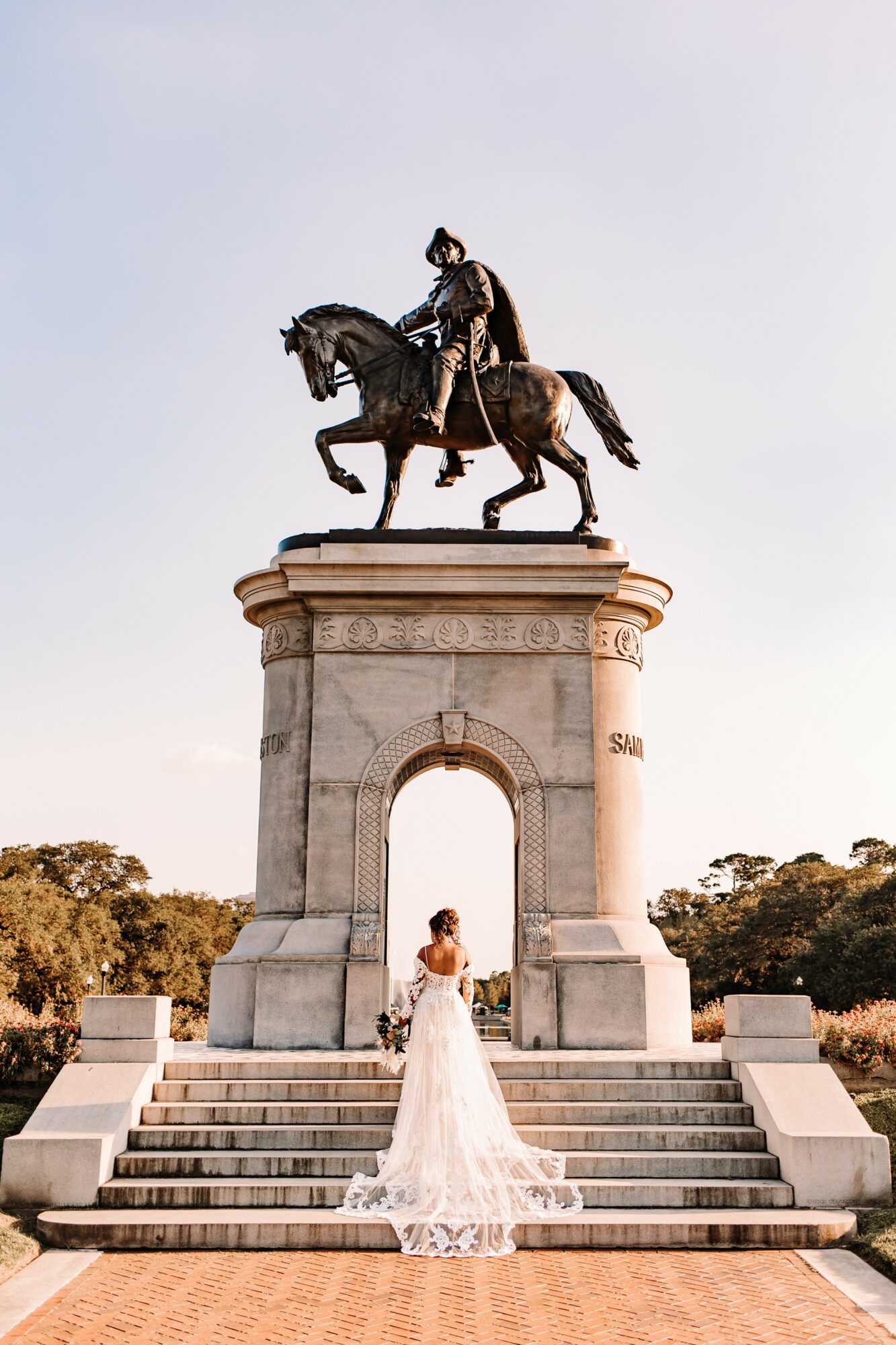 Woman in a white dress stands on steps beneath a large archway with a statue of a rider on a horse above, trees in background.