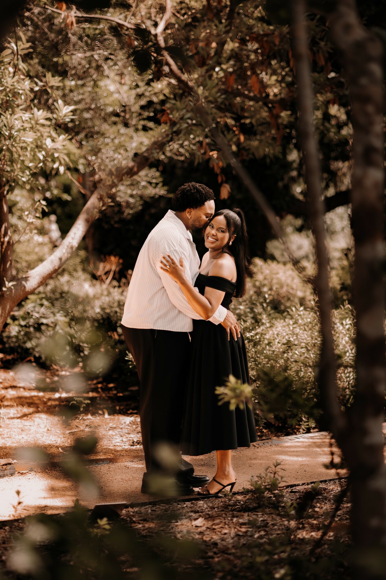 A couple stands close together in a wooded area, embracing and smiling at each other, surrounded by trees and sunlight.