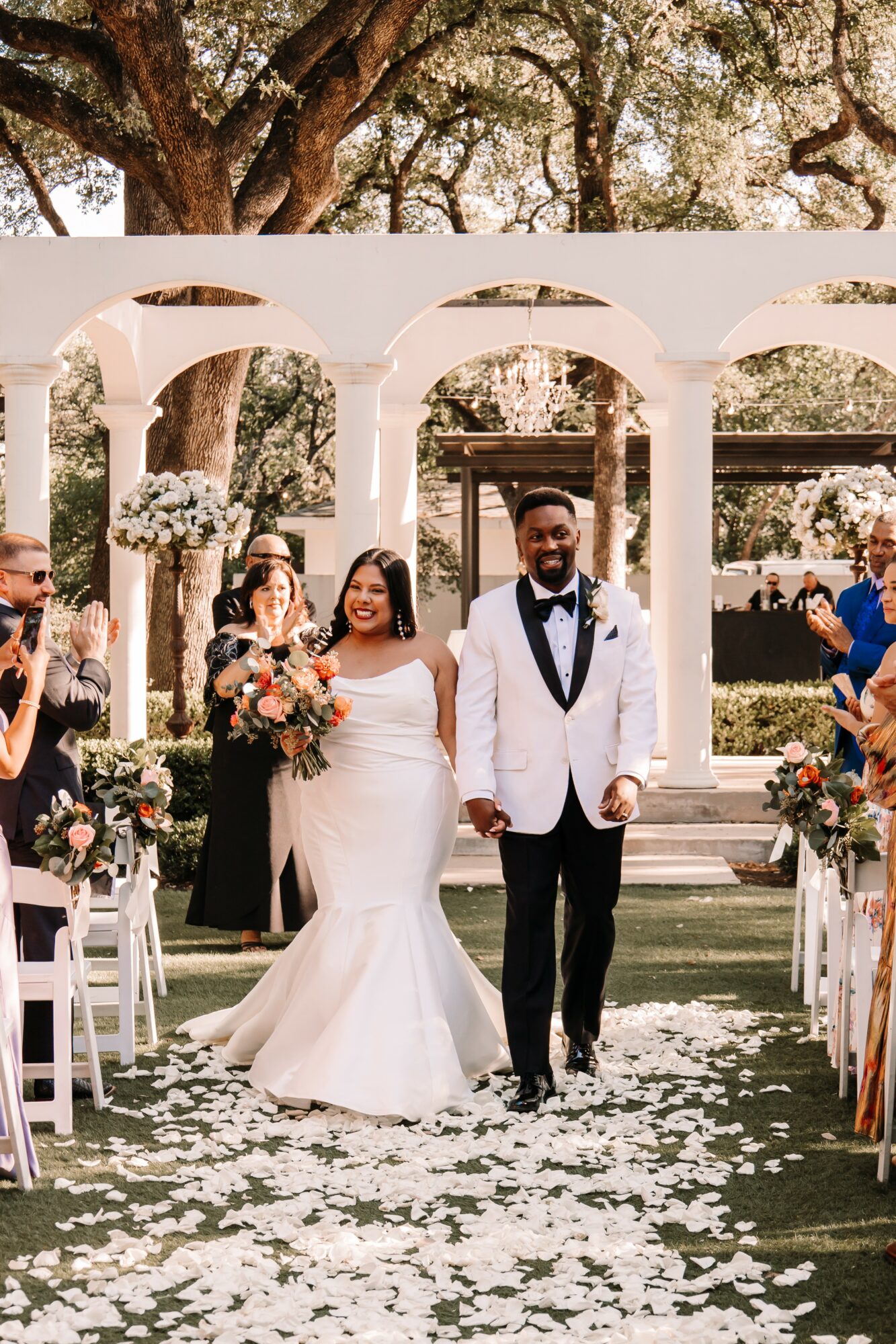 Bride and groom walking down the aisle at an outdoor wedding ceremony, surrounded by guests and floral decorations.