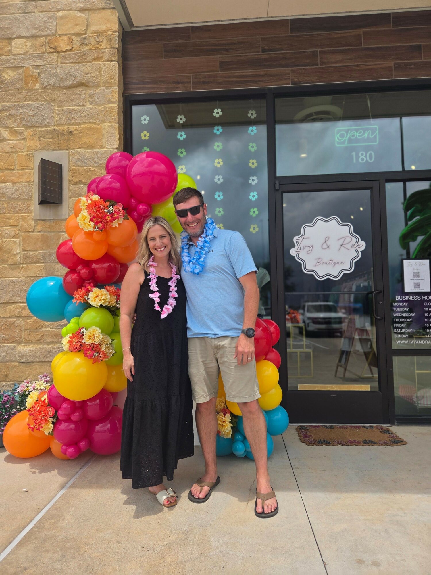 Two people dressed in costumes stand outside a building with balloons around them, one in a pink costume and the other in a black dress.