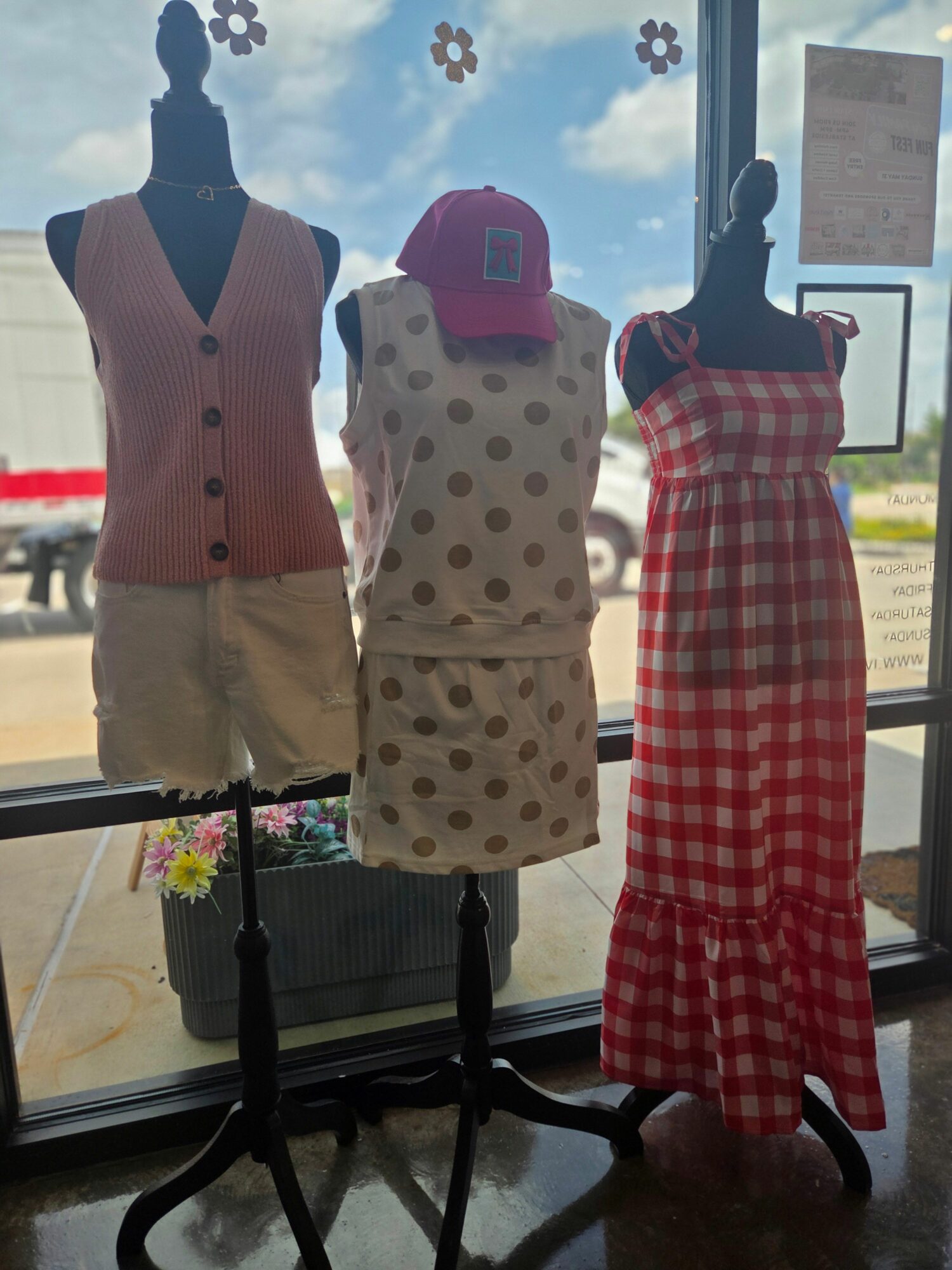 Three mannequins display clothing inside a store with a window showing a cloudy sky and outdoor scenery.