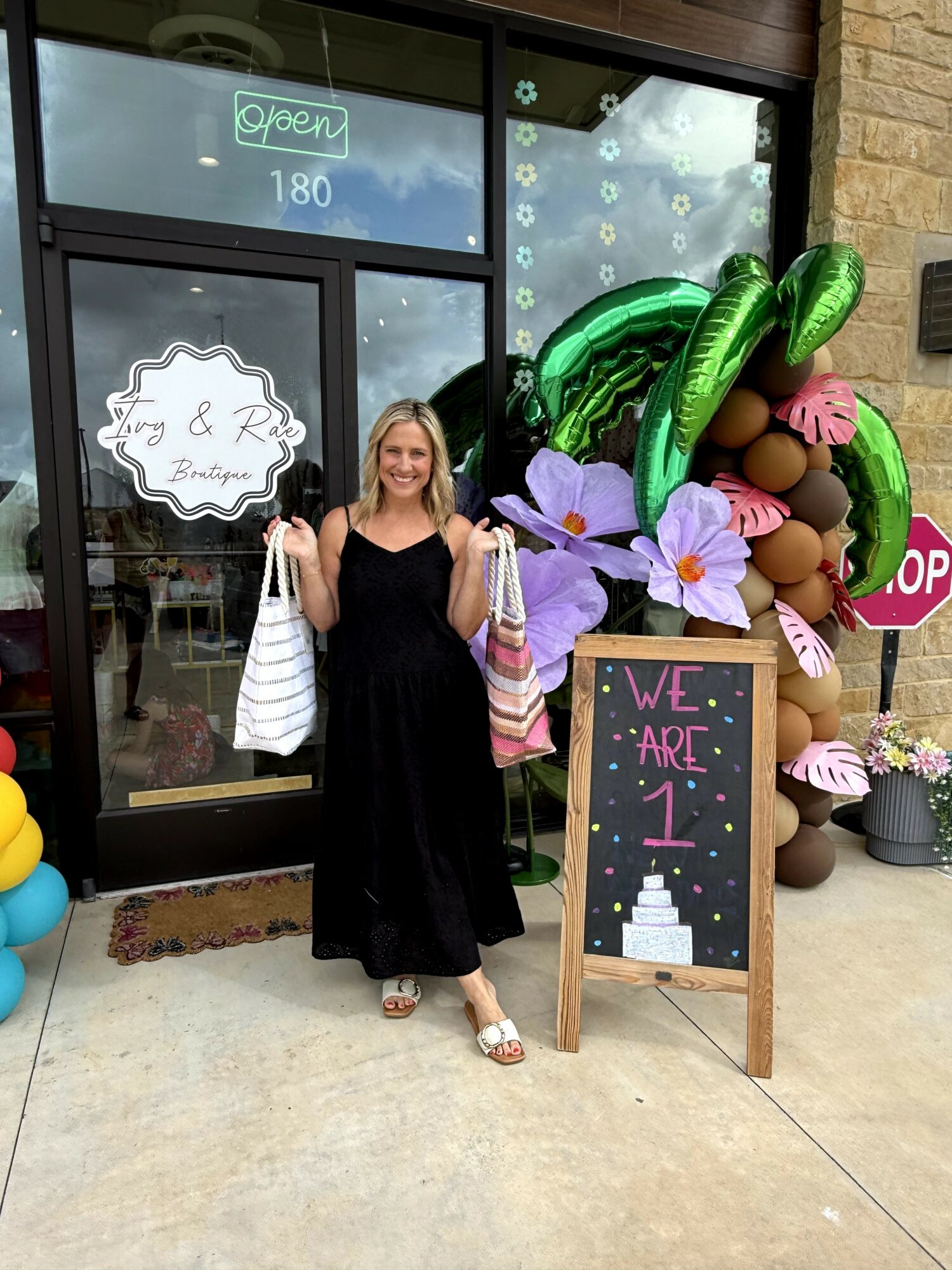 Woman standing outside a store with a chalkboard sign, balloons, and flowers, holding shopping bags, smiling.