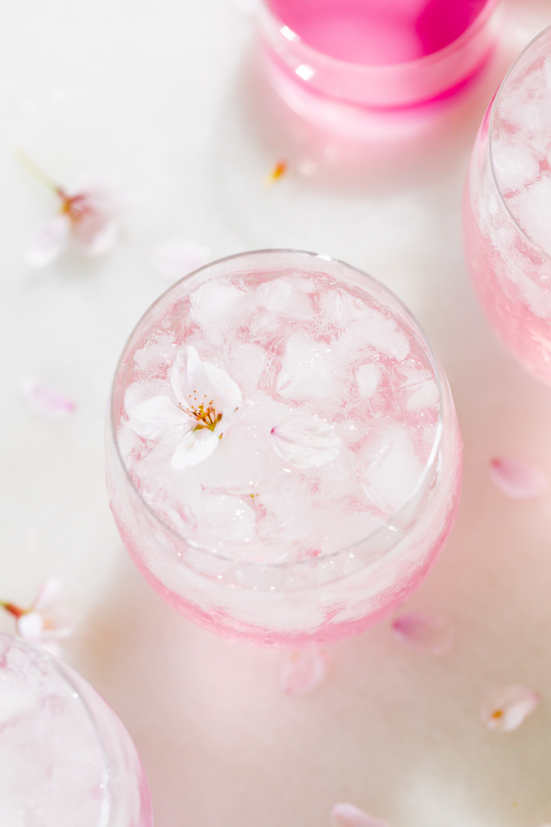Pink drink with ice in a glass, surrounded by flower petals and a pink container, on a white surface.