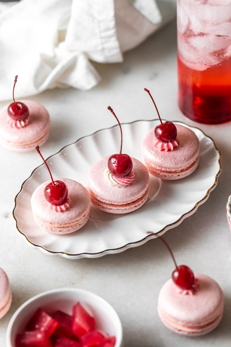 Pink macarons topped with cherries on a white plate, with a glass of red drink in the background.