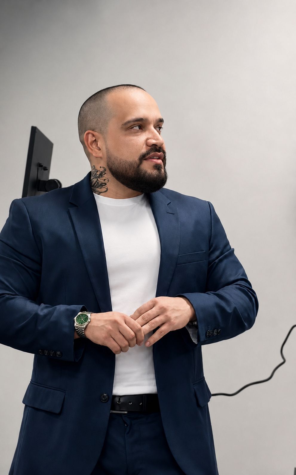 Man with a beard and tattoos wearing a suit, standing against a plain wall, looking to the side.