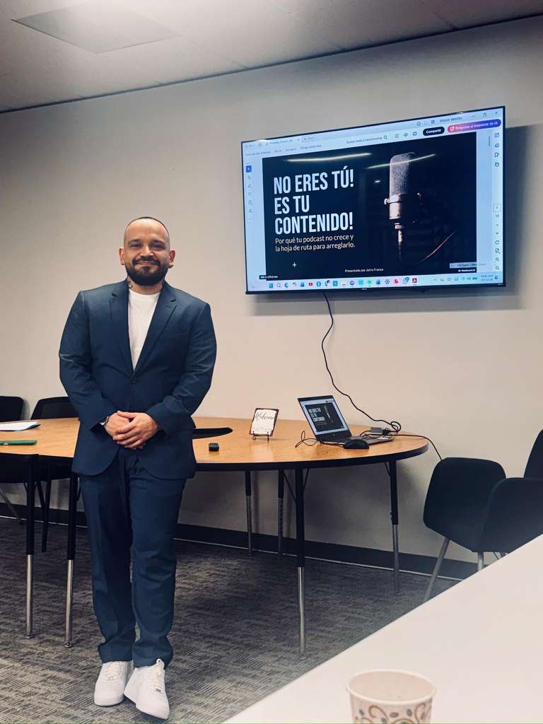 Man in suit standing in a conference room with a large screen displaying a message in Spanish, and a table with devices.
