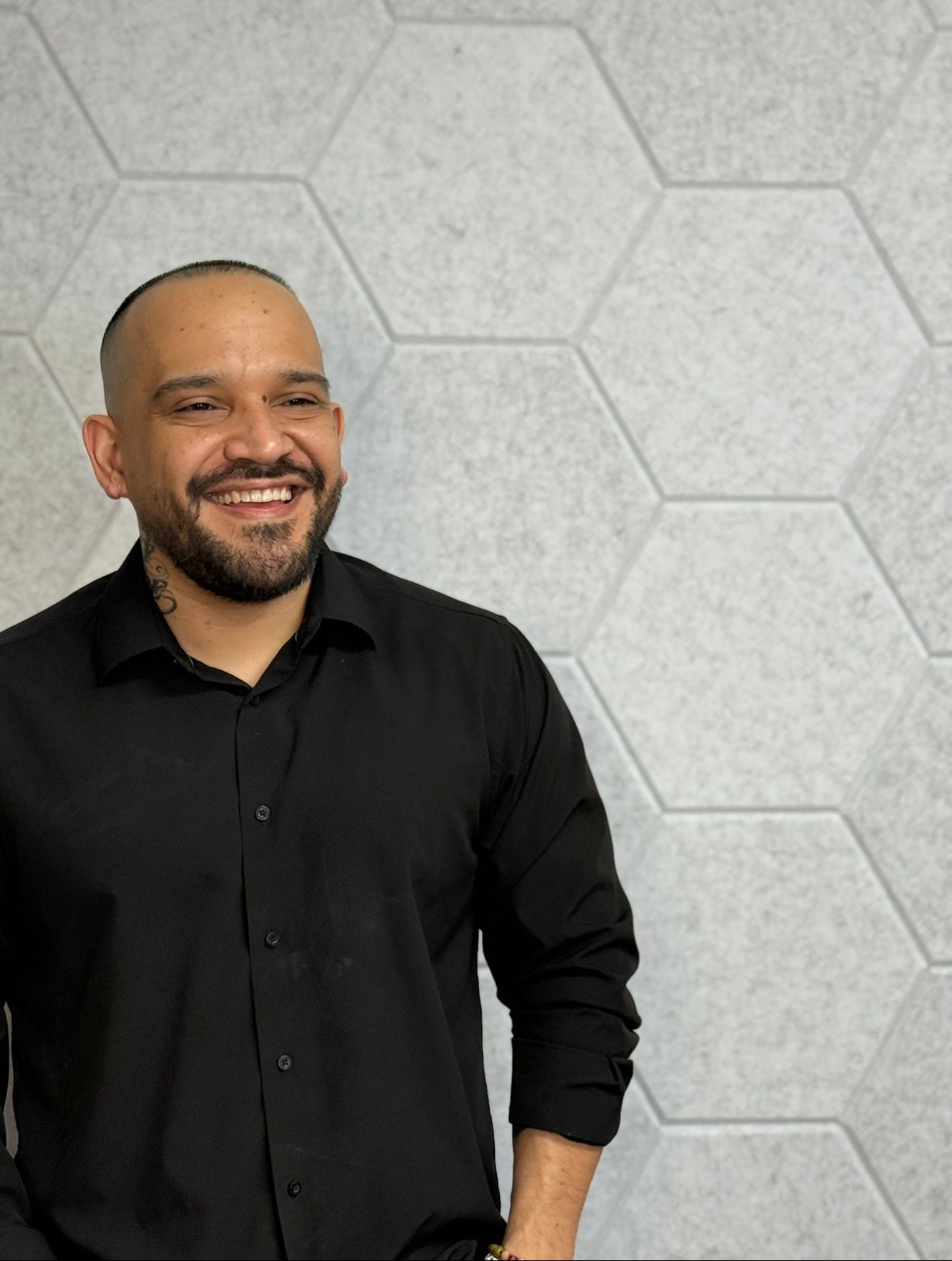 Smiling man with a beard and shaved head wearing a black shirt, standing against a hexagonal patterned wall.